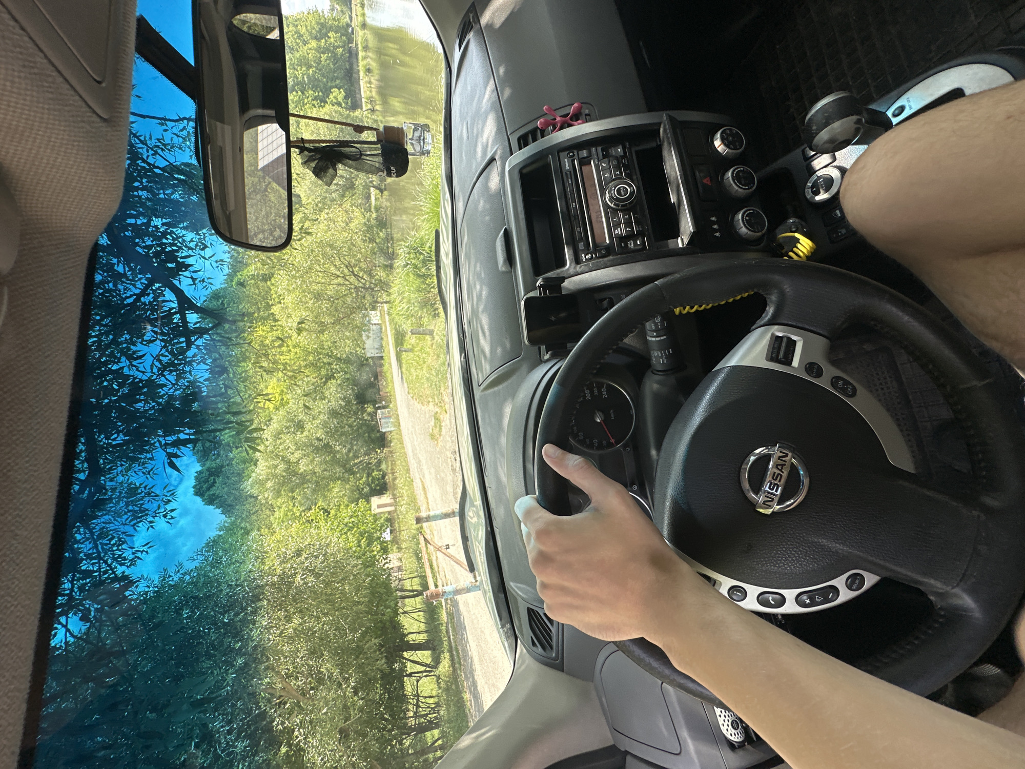 The image shows a first-person view from inside a Nissan vehicle parked on a dirt road. A person's left arm and hand are visible, resting on the steering wheel. The dashboard, steering wheel, and gear shift are prominent. Through the windshield and side windows, a body of water, trees, and a bright blue sky are visible. In the distance, there are what appear to be small buildings or structures. A car air freshener is hanging from the rearview mirror. The scene is consistent with a park or natural area, possibly a waterfront location. The presence of a dirt road suggests the vehicle is not in a typical urban setting. The context of the city of Makiv, Ukraine, is not directly evident from the visual cues alone but could be inferred from metadata or external information.