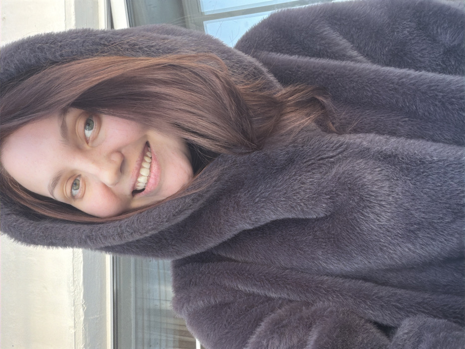 A young woman smiles directly at the camera, positioned at an angle. She is framed by a large, plush grey fur coat, with the hood pulled up around her head. Her brown hair peeks out from under the hood, cascading around her face. Her fair skin is highlighted by the light, emphasizing her green eyes. A window pane is visible in the background, indicating an indoor setting. The focus is sharp on her face and the texture of the fur coat.