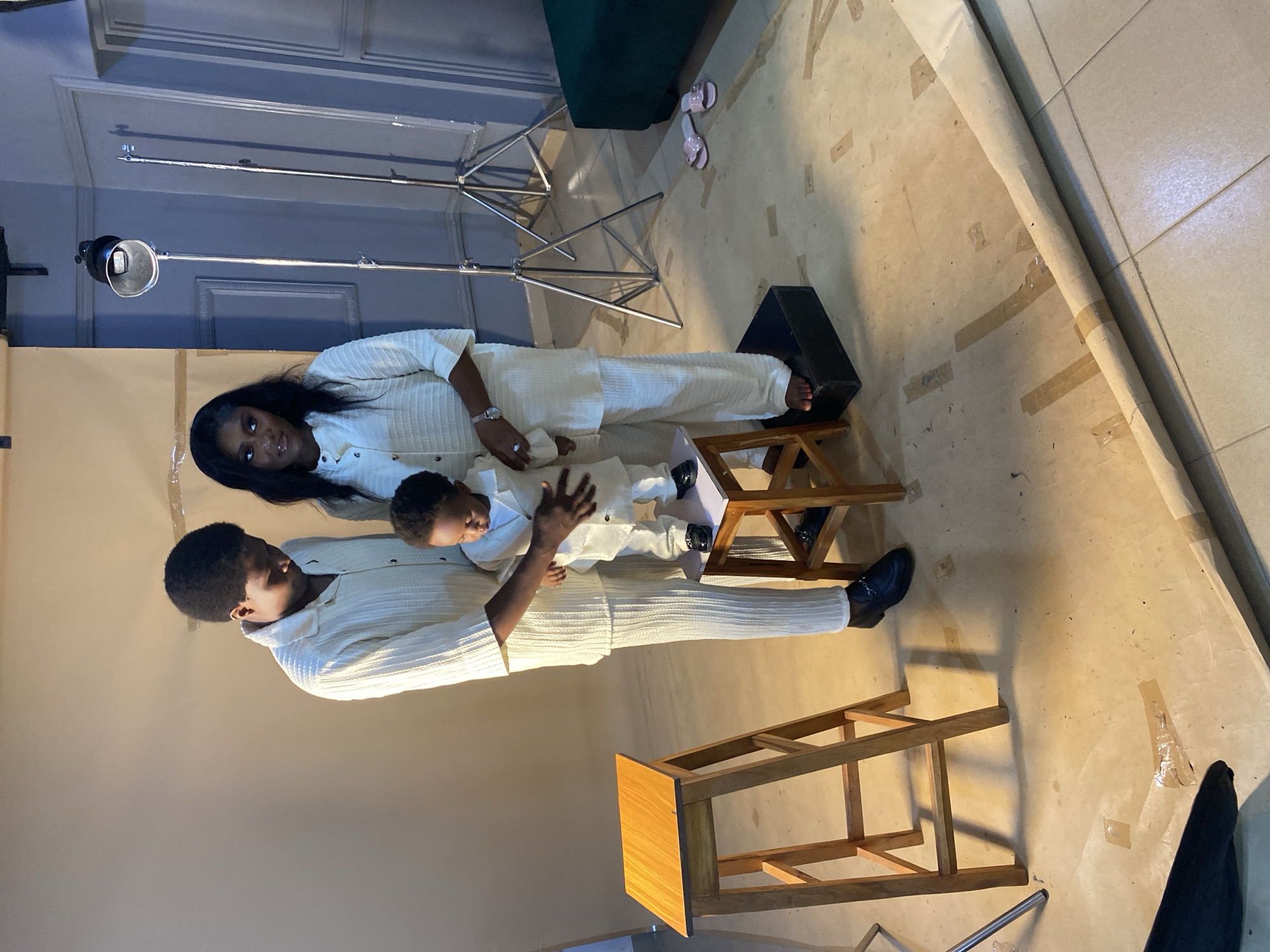 In a studio setting, a family of three is positioned on a light beige floor covered with a large roll of brown paper. To the left, a young boy stands on a small, square black platform, looking down at a toddler who is being held by a woman. The toddler, dressed in a white suit, is being supported by the woman's hands. The woman, with long dark hair, is wearing a matching white suit. To the right of the toddler, a man, also in a white suit, is placing his hand on the toddler's back. He has short dark hair and is looking down at the child. In the foreground, a wooden stool with a light brown top is visible. The background features studio lighting equipment, including two light stands with reflectors, and a dark green upholstered object. The walls are a muted blue with decorative molding. The overall scene suggests a professional photo shoot. The location is identified as Bukuru, Nigeria.