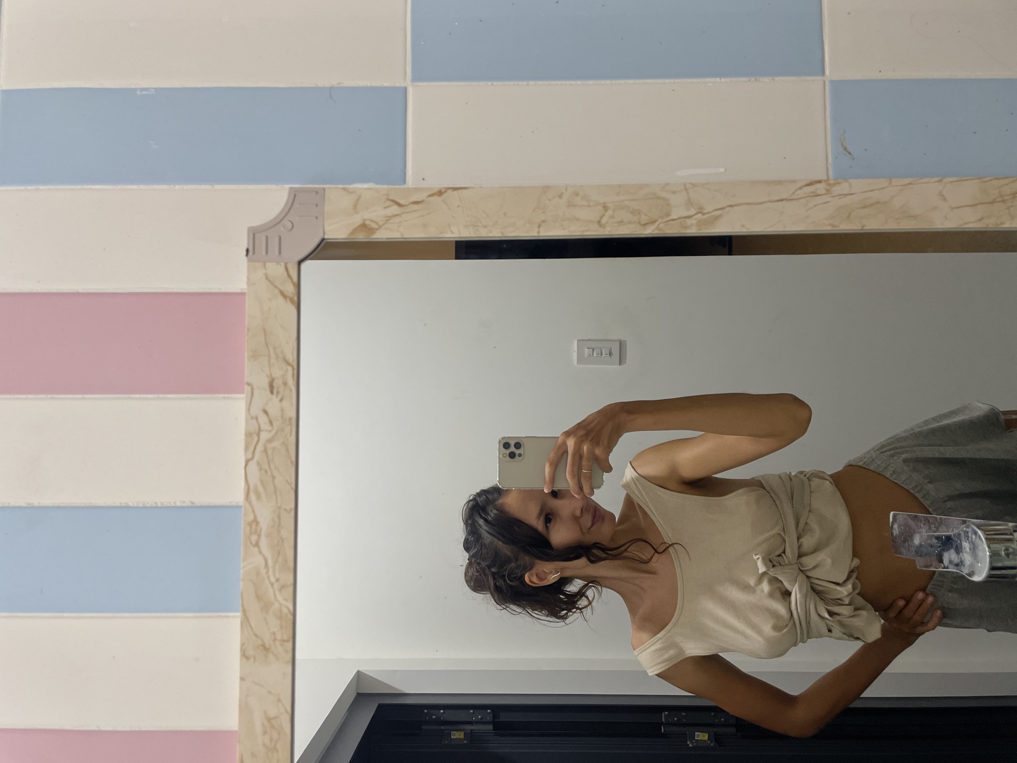 A woman is captured in a mirror selfie, standing in what appears to be a bathroom in Mũi Né, Vietnam. The image is rotated 90 degrees clockwise, showing the woman's reflection in a large mirror.
The woman has dark, curly hair tied back, and she is wearing a light beige, sleeveless top that is tied at the waist, exposing her midriff. She is also wearing grey, casual bottoms. She holds a light-colored smartphone (possibly an iPhone with multiple cameras) up to her face, angled towards the mirror to capture her reflection. A faint, pleasant smile is visible on her face. Her arms are slender, and a ring is visible on her left hand (right in the reflection) holding the phone.
The mirror has a decorative, marbled beige frame with a detailed, light-colored corner piece at the top right. Reflected in the mirror behind the woman is a white wall with a light switch featuring three horizontal rectangular buttons. Below the mirror's reflection, a dark surface, possibly part of a cabinet or doorway, is visible.
To the left and above the mirror, the wall is tiled with horizontal stripes. The tile pattern consists of alternating bands of white, light blue, and pink tiles, creating a soft, pastel aesthetic. Some minor scuffs or dirt marks are visible on the tiles. The lighting suggests an indoor setting, likely during the day, given the clear illumination. No discernible text is visible in the image.