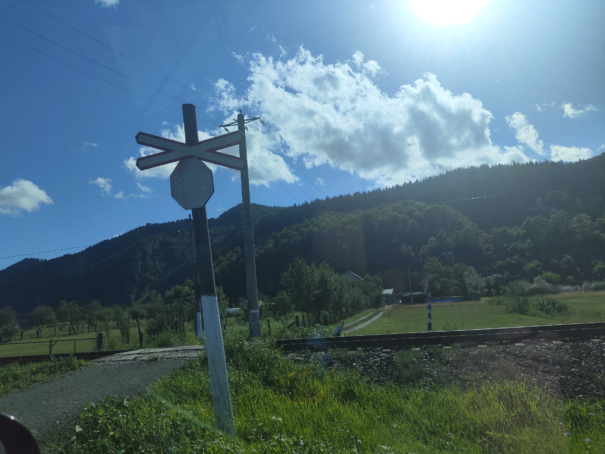 In the city of Leordina, Romania, under a bright sun and partly cloudy sky, a railway crossing sign stands prominently in the foreground. The sign, a post with a white and black striped base, features a white octagonal sign and a red and white "X" shaped crossbar above it. Adjacent to the sign, a single track railway line runs horizontally across the frame, laid on a bed of ballast. Beyond the tracks, a lush green field stretches towards a dense forest that covers a rolling hillside. A dirt road winds through the field, leading to a small building with a white roof. The environment is rural and natural, with power lines crisscrossing the sky.