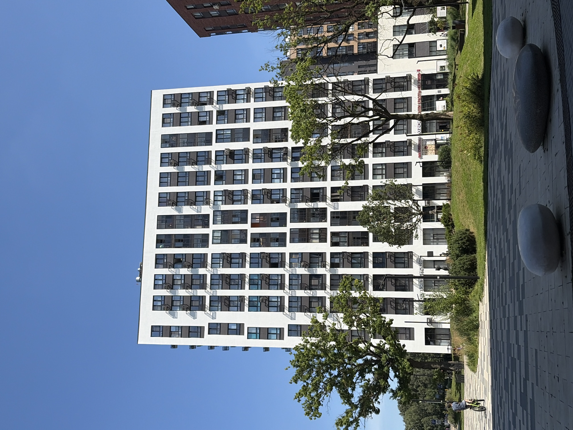In the city of Kommunarka, Russia, a modern, multi-story white apartment building dominates the frame under a clear blue sky. The building features a grid of dark-framed windows and balconies, with air conditioning units visible on some. To the right, a partially visible brick building stands as a contrast. In the foreground, a paved walkway with a geometric pattern leads to a grassy area with scattered trees and bushes. A person on a yellow bicycle is seen in the mid-ground, riding on the walkway. Large, smooth, grey and white decorative stones are placed on the paved area in the foreground. The scene depicts a contemporary urban residential environment with outdoor recreational space.