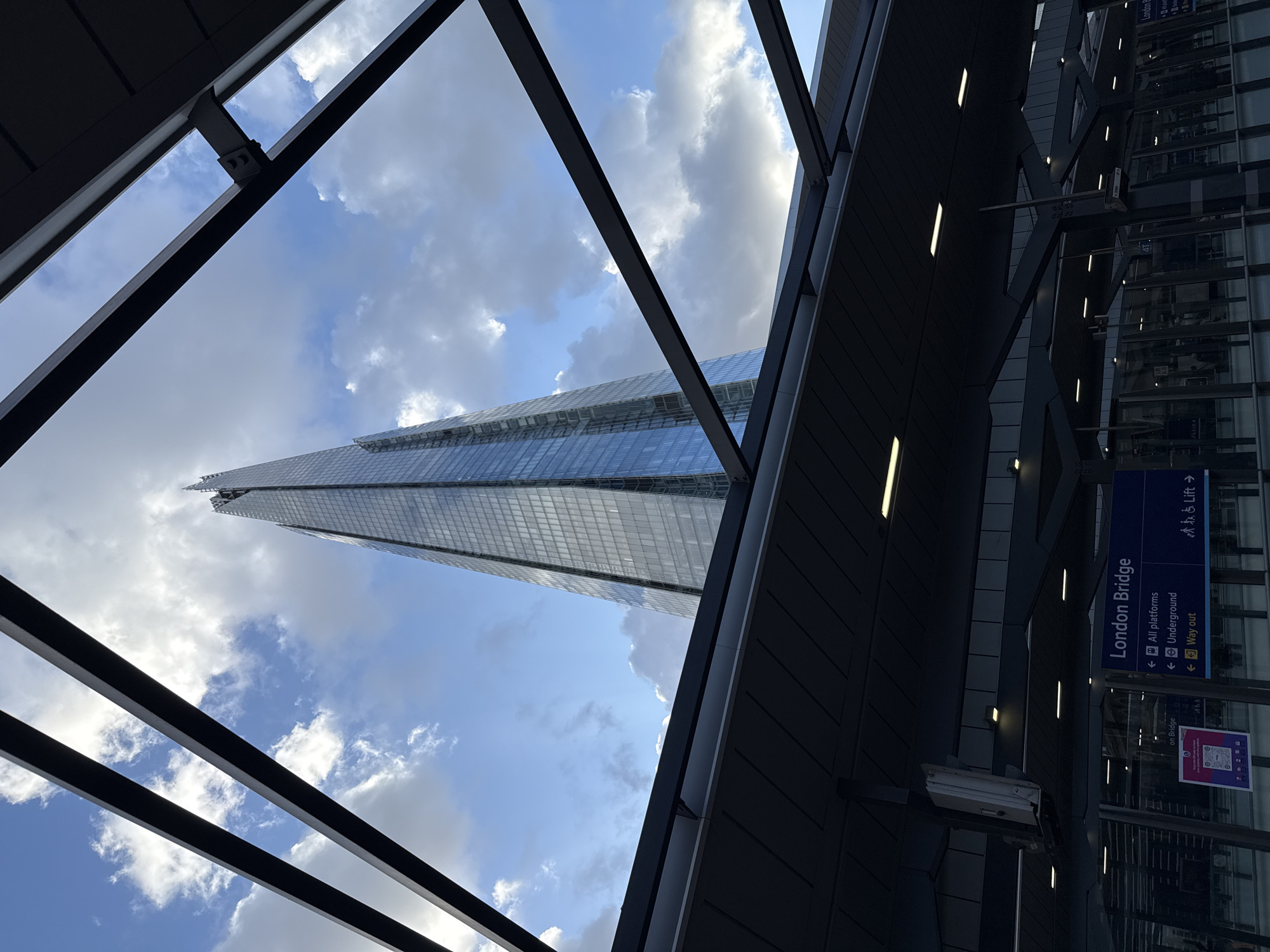 The image presents a view of The Shard skyscraper in London, United Kingdom, framed by the structural elements of what appears to be the London Bridge railway station. The Shard, with its distinctive tapering shape and glass facade, dominates the sky, visible between dark metal beams and supports. The sky itself is partly cloudy.
To the right, a sign indicates "London Bridge" with directions for "All platforms," "Underground," and "Way out," along with a symbol for "Lift." The scene is urban, emphasizing architecture and transport infrastructure.
