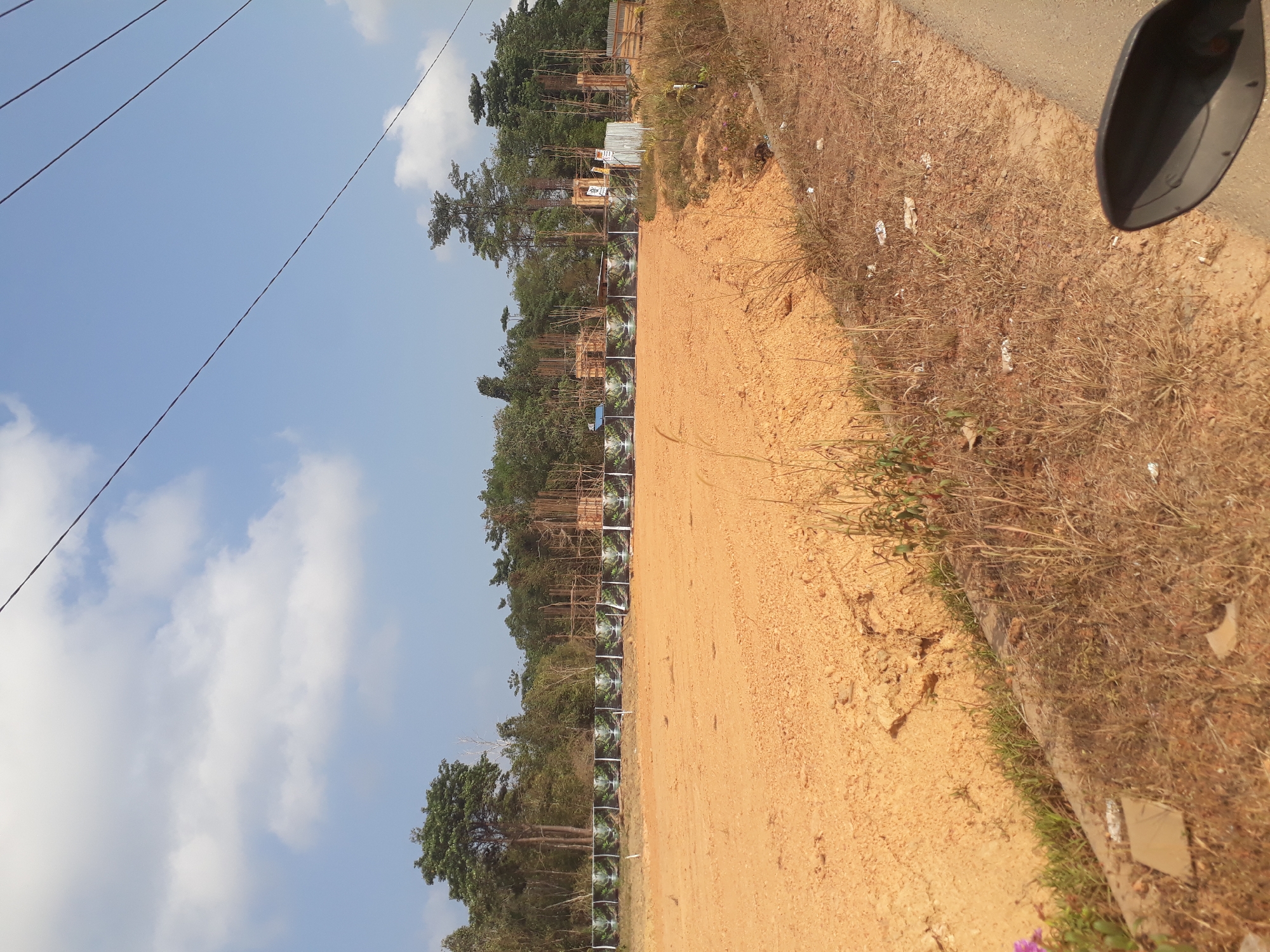 The image depicts an undeveloped construction site in Desa Balun Ijuk, Indonesia, under a bright blue sky with scattered white clouds. The foreground is dominated by a large area of orange-brown soil, likely prepared for construction, with tire tracks visible on its surface. A low concrete barrier separates this area from a strip of dry grass and weeds, with some scattered debris. In the background, a row of large, dark banners featuring images of waterfalls and lush greenery stretches across the site. Beyond the banners, trees form a dense backdrop. Visible construction elements include scaffolding and rebar structures, indicating ongoing building activity. Power lines crisscross the upper portion of the image. The scene suggests a new development project in its early stages, with elements of natural landscaping represented in the banners contrasting with the raw construction environment.