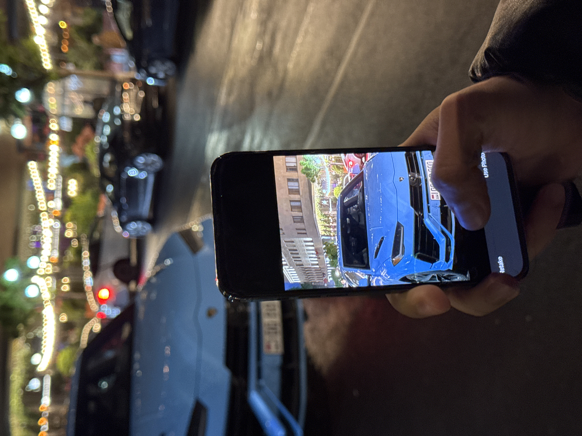 A person is using a smartphone to take a picture of a light blue sports car parked on a street in Yerevan, Armenia. The car being photographed is the same light blue as the car the person is standing next to. The original image on the smartphone is in portrait mode and shows a front view of the light blue sports car parked in front of a building. The environment is an urban setting at night, with streetlights visible in the background, including some bokeh orbs of light to the left of the smartphone.