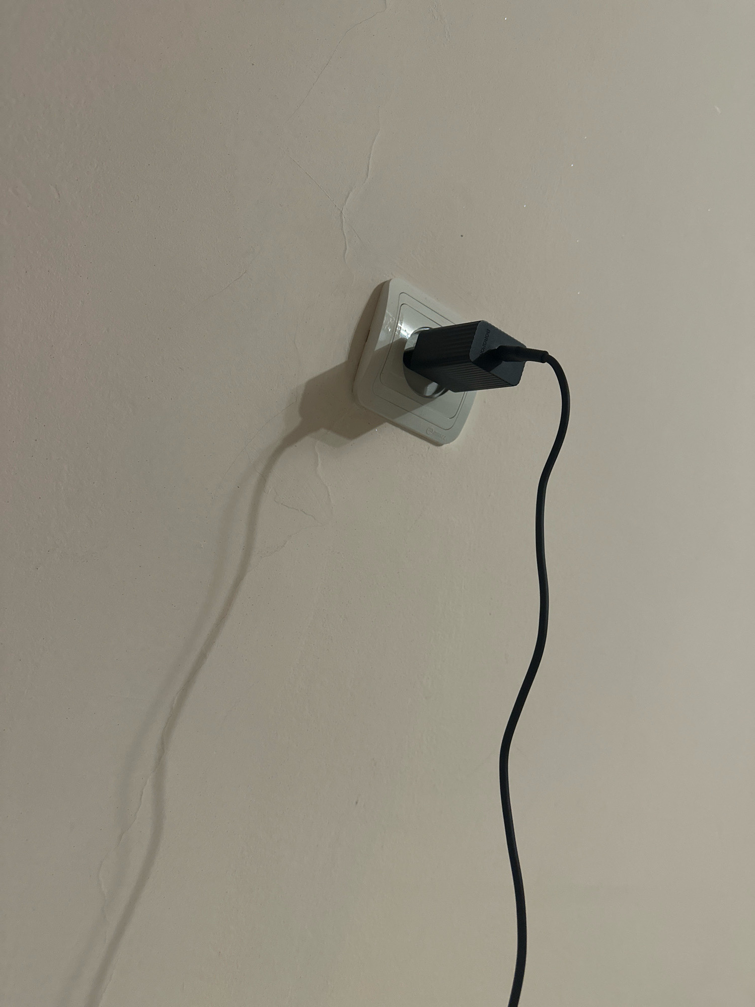 An electrical outlet is mounted on a beige wall. A black electrical adapter is plugged into the outlet, and a black cable extends from the adapter. The cable casts a long shadow on the wall. The wall shows signs of minor cracks and texture variations. The scene is indoors. The location is not specified in the city of Gyumri, Armenia.