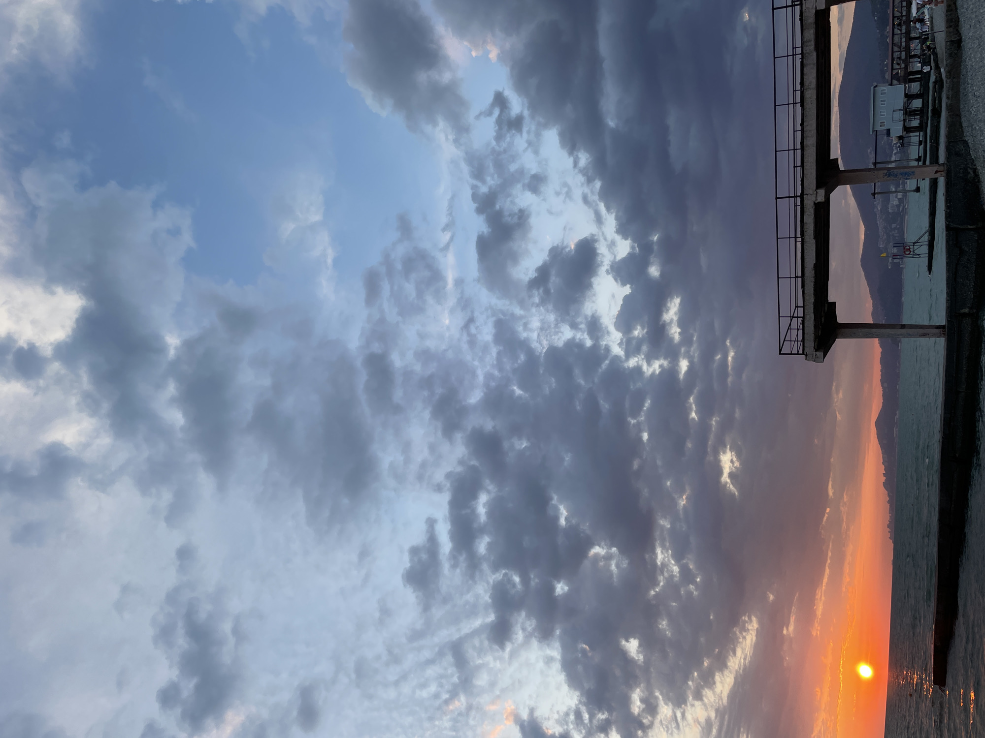 This is a vertical, eye-level, medium shot of the Black Sea coast in Sochi, Russia, at sunset. The sun is a bright orange orb low on the horizon to the left, casting a warm glow on the water and the sky. Dark, heavy clouds dominate the upper portion of the frame, with patches of lighter blue sky peeking through. In the foreground, a concrete pier extends from the right side of the image into the sea, with a black metal railing along its edge. The pier is supported by concrete pillars. Further in the distance, a line of mountains can be seen against the horizon, with buildings and structures scattered along their slopes and at their base. Some people are visible on the pier and on the shore in the distance, though they are small and indistinct. A small, light blue building with white windows is located on the pier in the mid-ground. The water is a deep blue-green, with small waves. The overall atmosphere is one of a dramatic and cloudy sunset over a coastal city.