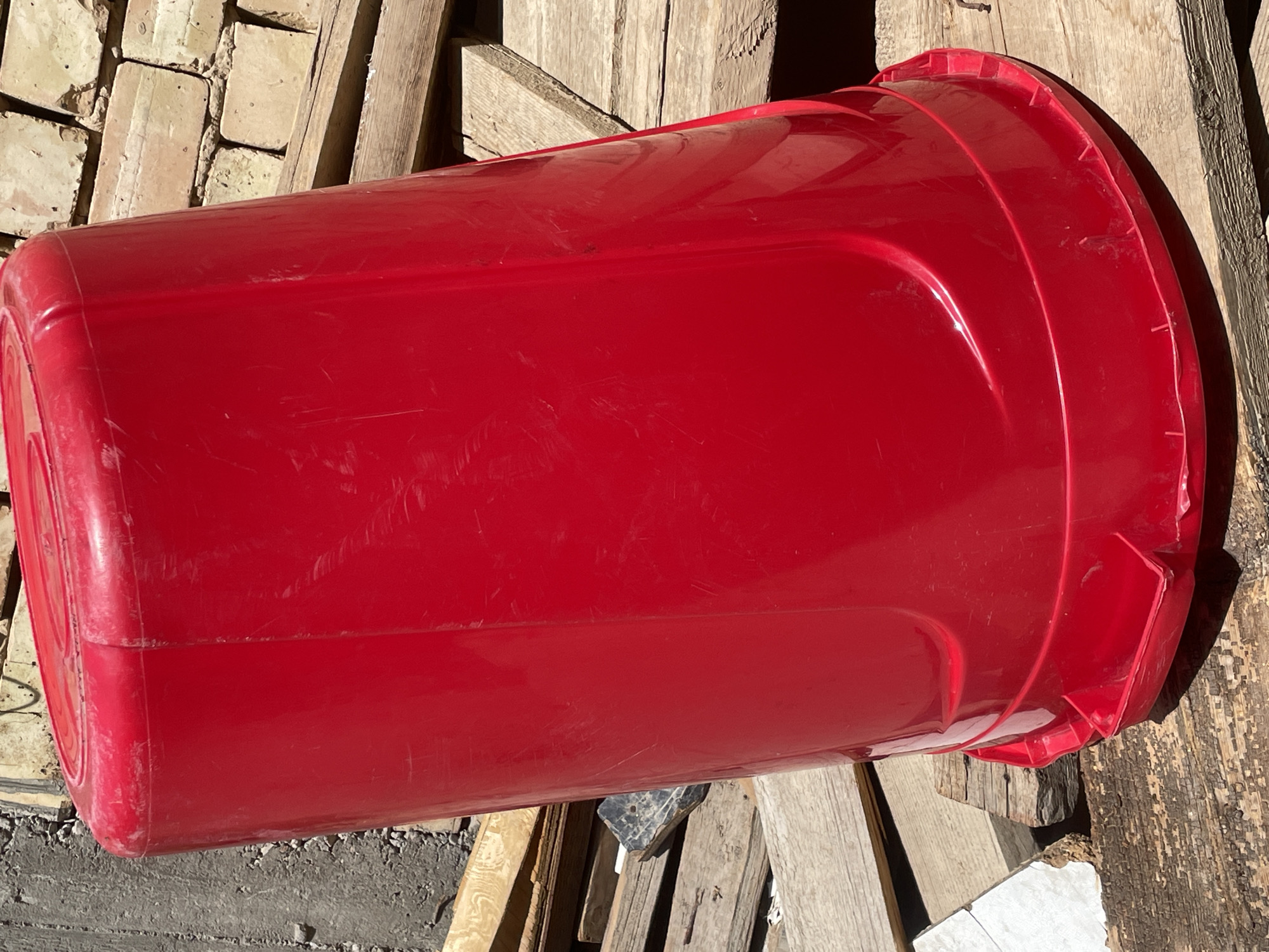 A large, bright red plastic bucket is the primary subject, positioned upright and slightly tilted. The bucket has a rounded lid and a wide base with a reinforced rim. Its surface shows signs of use, with minor scratches and scuffs. The bucket is resting on a collection of weathered wooden planks, some of which appear to be pallet wood. Behind the bucket and planks, a textured gray concrete wall is visible on the left, and a yellowish brick wall is seen on the right. The scene is outdoors, illuminated by bright sunlight, casting distinct shadows. The overall environment suggests a construction site, a workshop, or an area where materials are stored or processed. The image was taken at an outdoor location in Tashkent, Uzbekistan.