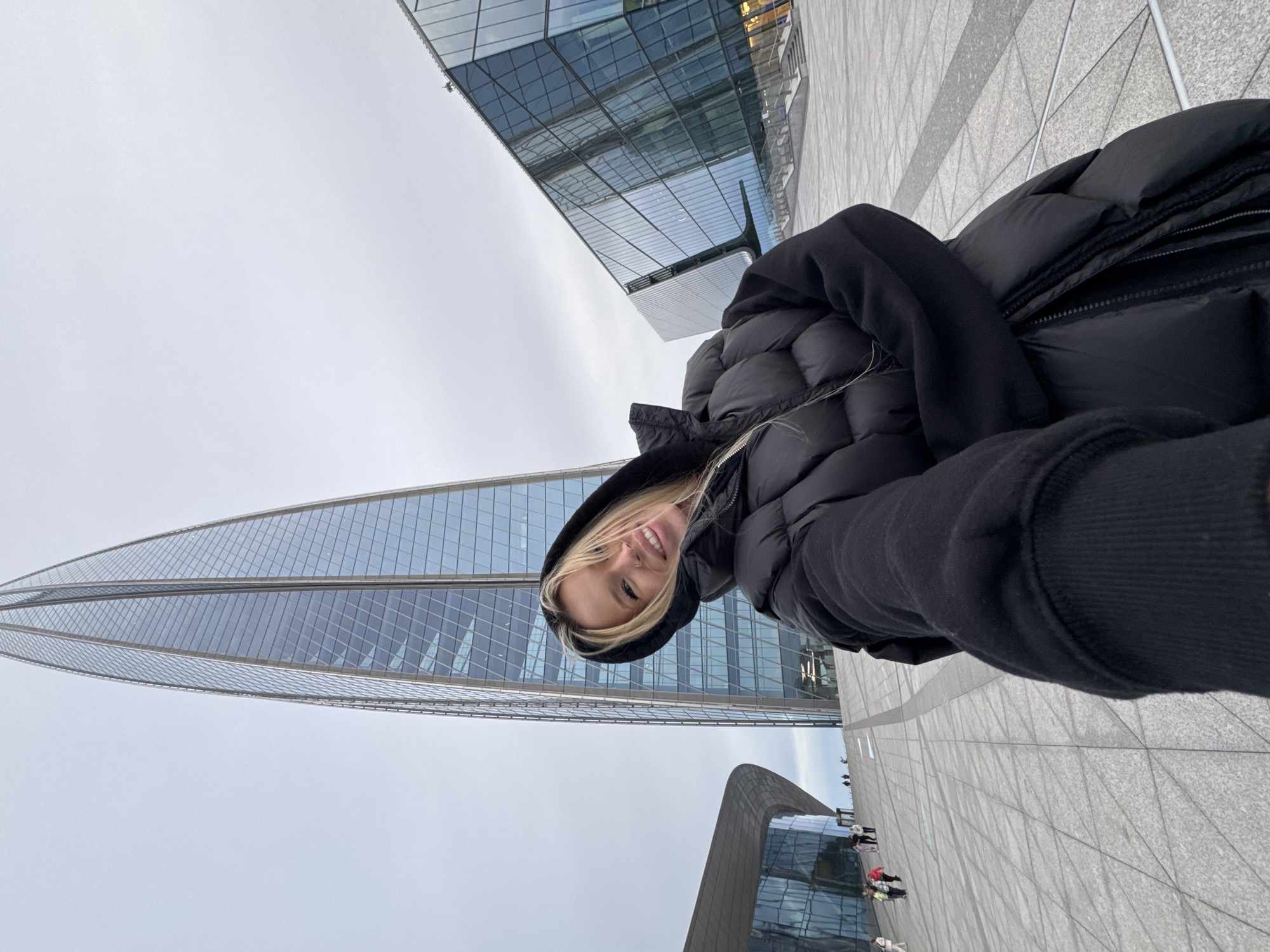 The image shows a young woman with blonde hair wearing a black puffer jacket and a black hoodie. She is smiling at the camera, suggesting a cheerful or happy emotion. The photo appears to be a selfie, taken from a low angle.
The setting is an outdoor urban environment with modern architecture. In the background, there are several large buildings with glass facades. One prominent building is a tall, sleek skyscraper with a curved, twisted design, reflecting the overcast sky. Another building in the foreground has a dark, undulating roofline. The ground is paved with large, grey tiles arranged in a grid pattern.
The weather appears to be overcast or cloudy, indicated by the dull, diffused light and the grey sky. There are a few other people visible in the distance, walking on the paved area, but they are small and indistinct. The overall scene suggests a modern city location, likely during the daytime due to the ambient light.
The image is oriented with the woman's head towards the bottom left and her body extending towards the top right. This unusual orientation might be due to the selfie being taken at an angle. The clothing suggests cooler weather, consistent with a European city like Saint Petersburg. No text is visible in the image.