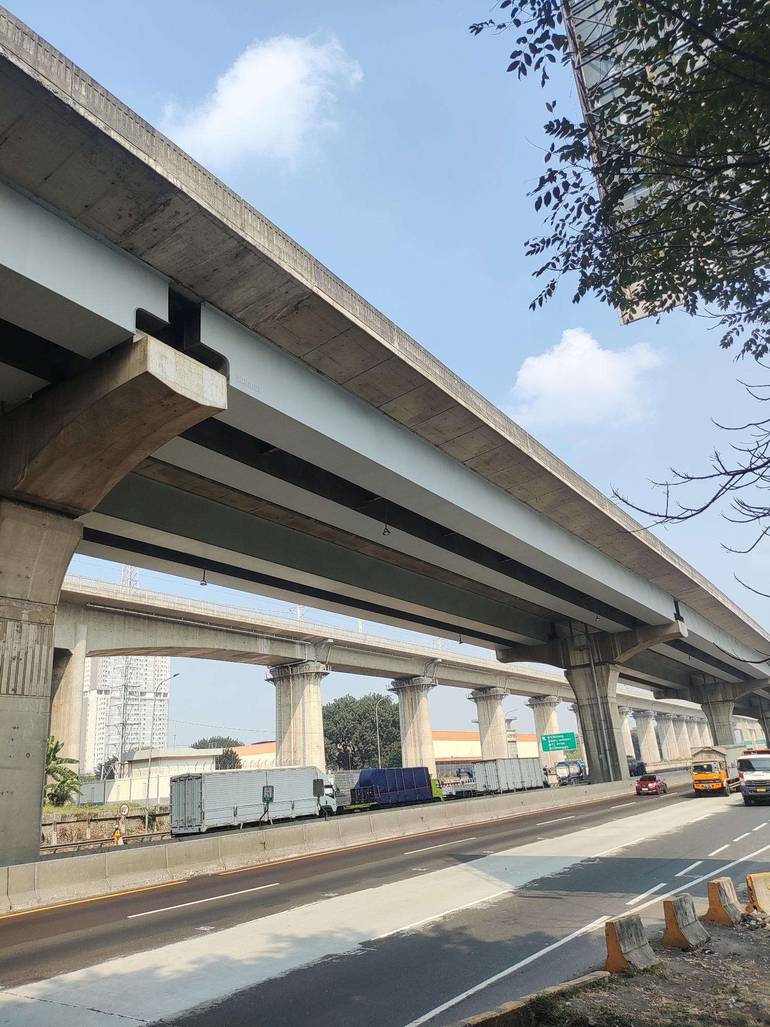 The image shows a multi-level highway overpass in Lambangsari, Indonesia. The sky is blue with scattered clouds. Several concrete pillars support the elevated roadways. On the lower level of the highway, trucks and cars are driving. One truck is white with a large container, and another is blue with a tarp covering its load. There are also orange and white traffic cones and barriers visible along the side of the road. In the background, there are tall buildings and trees.