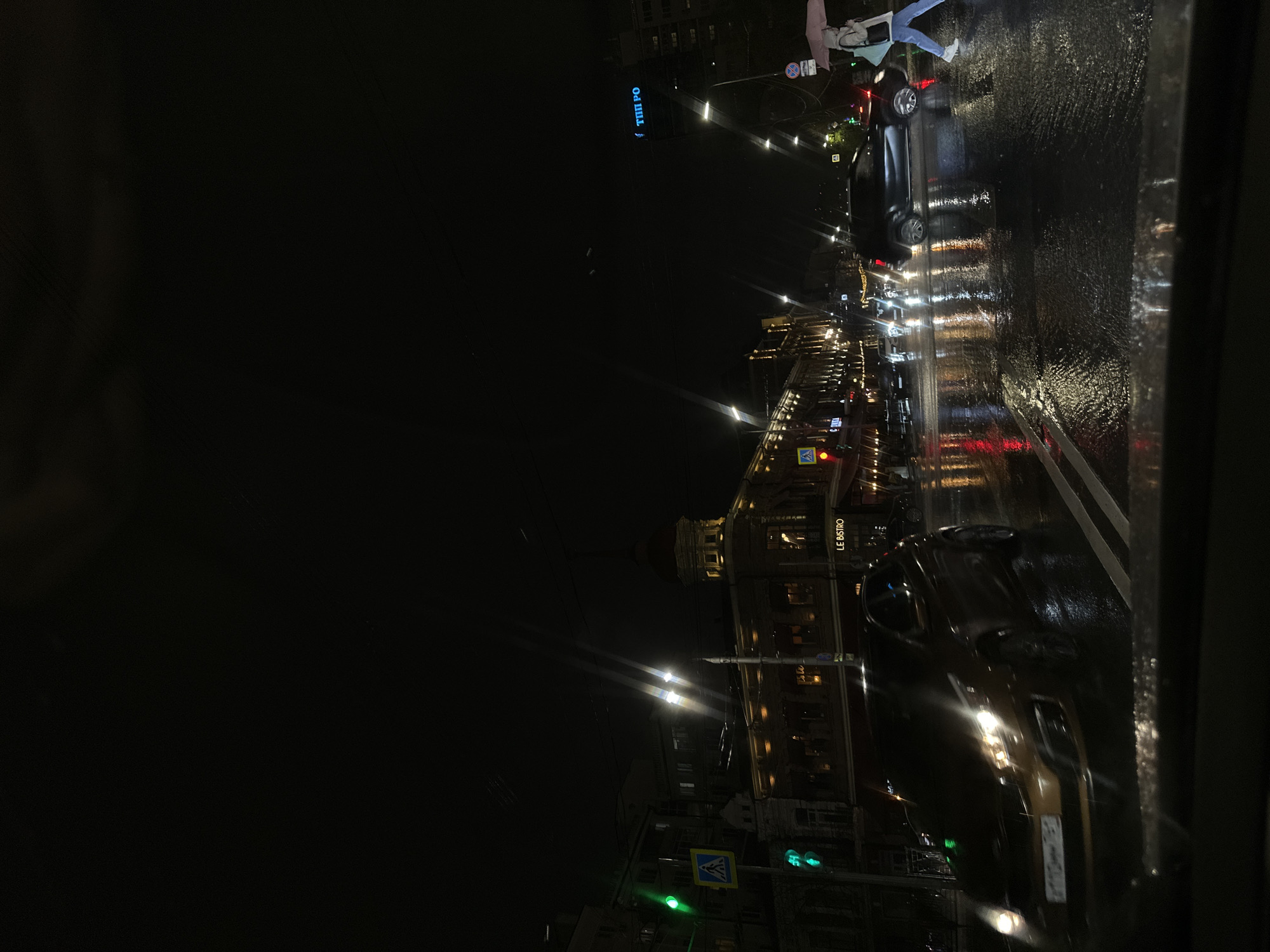 The image depicts a nighttime street scene in Ovoshchnoy, Russia. It appears to be raining, as the street is wet and reflects the city lights. There are several cars on the road, some with their headlights on. Buildings line the street, including one with the signage "LE BISTRO." Streetlights illuminate the scene, and traffic lights are visible. A pedestrian is crossing the street in the upper part of the image. The environment is dark, wet, and urban.