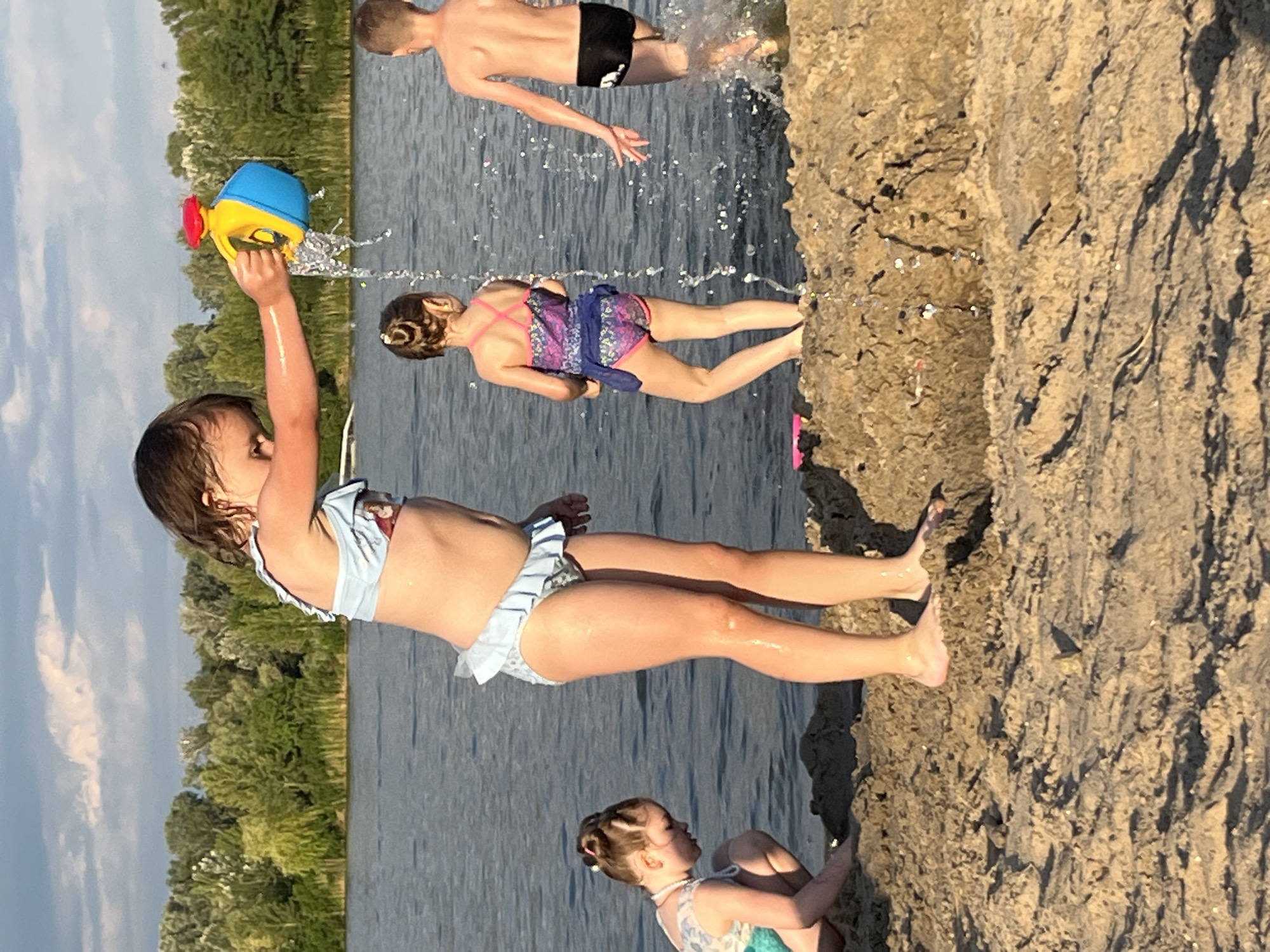 The image shows a group of children playing in the water near a rocky shore. A young girl in the foreground, wearing a light blue swimsuit, is holding a yellow and blue watering can, splashing water. Behind her, another girl in a purple swimsuit is partially visible. Further back, a boy in black swim trunks is jumping into the water, creating a splash. At the bottom right, a younger child with light brown hair is sitting on the rocks, wearing a swimsuit with a floral pattern. The scene appears to be set during a sunny day, possibly late afternoon, given the warm lighting and shadows on the rocks. The background shows a body of water, a line of green trees, and a cloudy sky. The overall atmosphere is one of playful enjoyment and summer recreation. The image is taken from a slightly elevated perspective, looking down towards the water and the people.