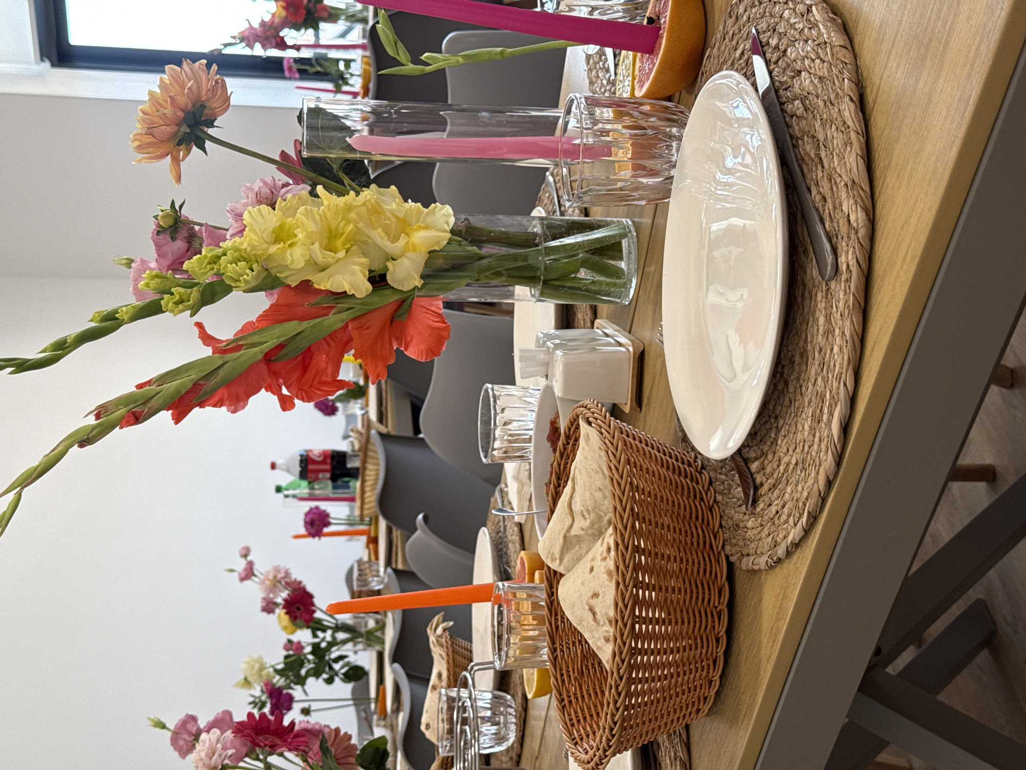 A table is set for a meal with floral arrangements and bottles of Coca-Cola and Sprite. The table is made of light-colored wood, and a light gray metal frame is visible underneath. The chairs around the table are dark gray. In the foreground, a woven basket contains flatbreads. There are white plates, glassware, and cutlery on the table. Candles in various colors are placed in decorative holders. A grapefruit cut in half is also visible on the table. The scene appears to be indoors, possibly in a dining room or restaurant. The lighting suggests it is daytime.