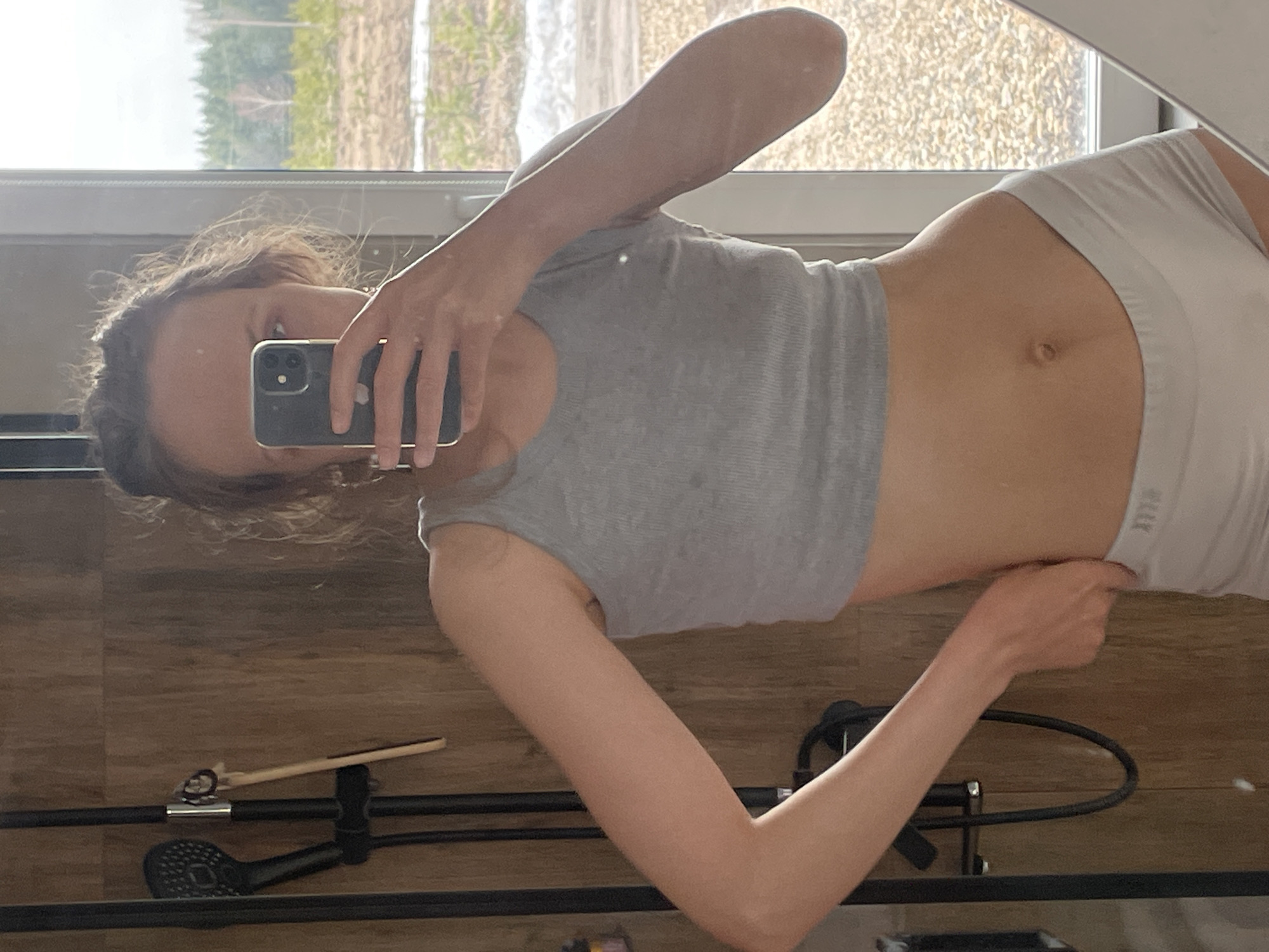 A woman is taking a selfie in a bathroom. She is wearing a gray crop top and white shorts. The background shows a window with trees outside and a shower head. The bathroom has wood-paneled walls. The location is potentially Подшивалово, Russia.