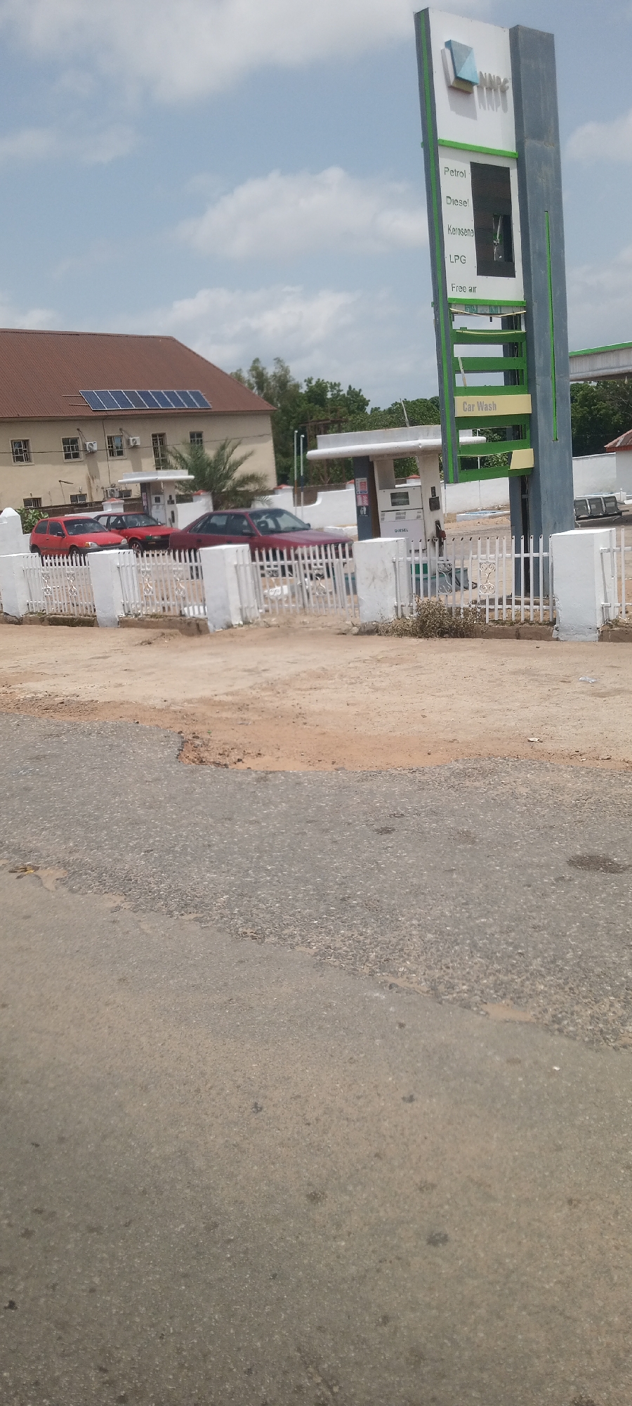 The image shows a gas station with a tall sign displaying "NNPC" at the top. Below the company name, various fuel types are listed: Petrol, Diesel, Kerosene, and LPG. "Free air" is also indicated, and further down, "Car Wash". Several cars are parked in front of the gas station, some red and one a darker maroon color. A white picket fence separates the street from the gas station area. Behind the cars, a building with a brown roof and solar panels is visible, along with air conditioning units. The sky is partly cloudy, suggesting it is daytime. The overall scene depicts a functional gas station, likely in a town or city setting, with vehicles and a building present. There are no people visible in the image. The ground in the foreground is a dirt and asphalt road, showing some signs of wear.