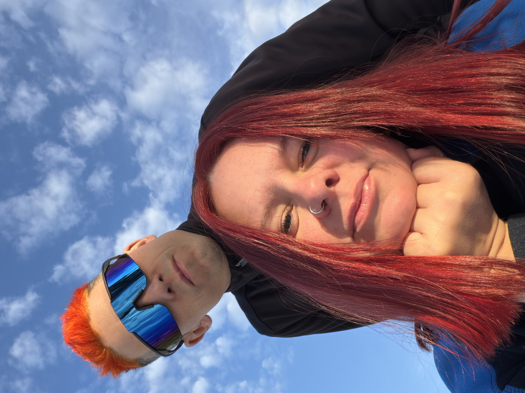 A low-angle, selfie-style photograph shows a woman with long, red hair resting her chin on her hand. Adjacent to her and also facing the camera is a man wearing reflective blue sunglasses and having short, orange hair. The background is a bright blue sky dotted with white clouds. The setting appears to be outdoors. There is no information in the image to indicate that it was taken in the city of Chaikovsky, Russia.
