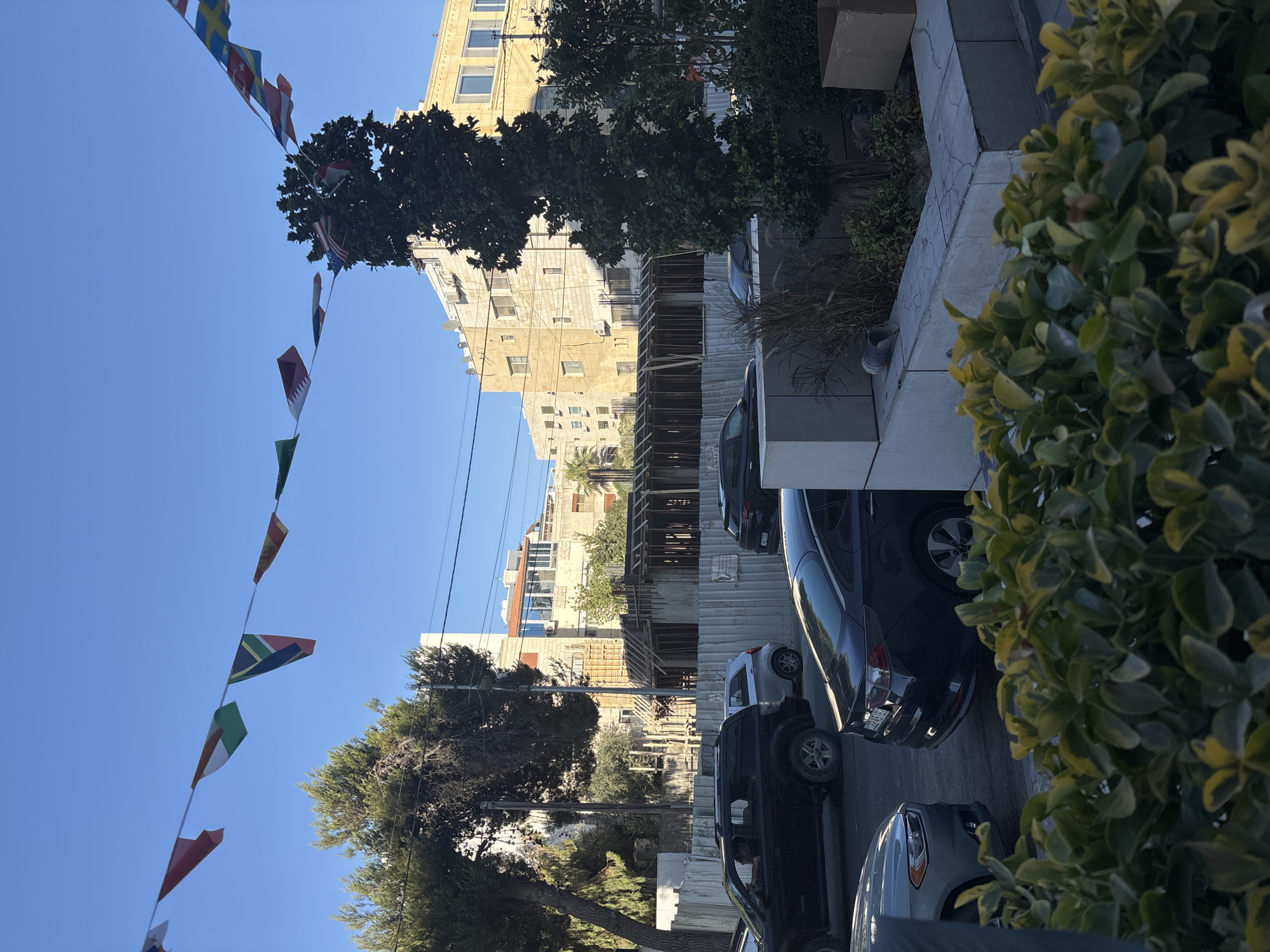The image captures a street scene in Amman, Jordan, under a clear blue sky. A string of international flags is strung across the upper portion of the frame. In the middle ground, construction is underway on a building, with wooden scaffolding and concrete structures visible. Several cars are parked or driving on the street, including a black SUV, a silver compact car, and a black sedan. Trees and buildings line the street, with some of the buildings appearing to be older, multi-story structures. A section of green foliage is in the foreground, partially obscuring the view of the street and construction site. The scene appears to be a typical urban environment with elements of ongoing development.