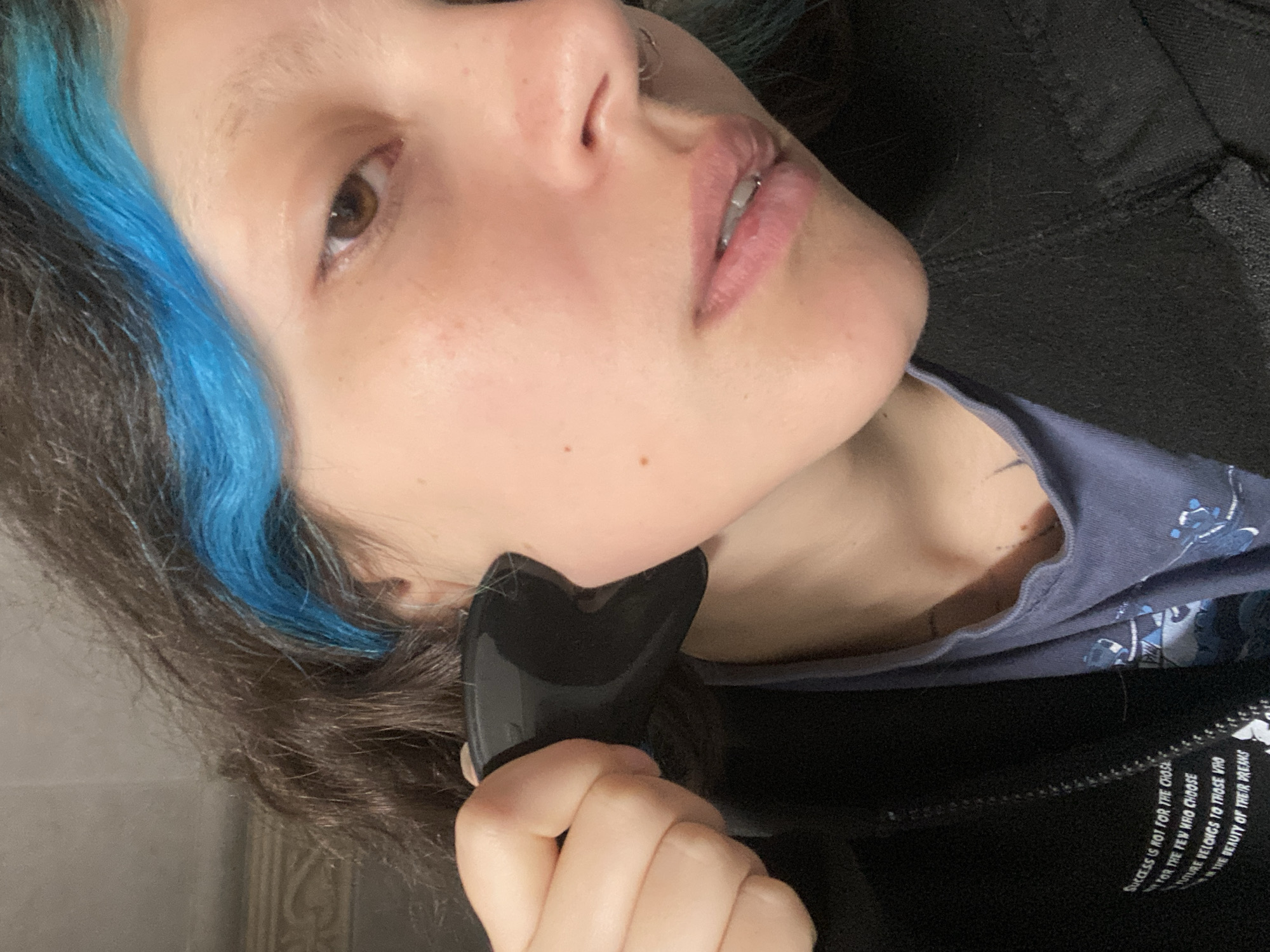 A close-up, slightly angled portrait shows a person with light skin, brown hair with a blue streak, and brown eyes. The person is using a dark, glossy gua sha tool on their face, specifically near the jawline. The individual is wearing a dark jacket over a blue t-shirt with a design. The image also captures a portion of text on the jacket, which reads "SUCCESS IS NOT FOR THE CHOSE / FOR THE FEW WRO CHOOSE / MTURE BELONGS TO THOSE WHO / THE BEAUTY OF THEIR DREAMS".
The location in the city of Gijón/Xixón, Spain, cannot be determined from this image.