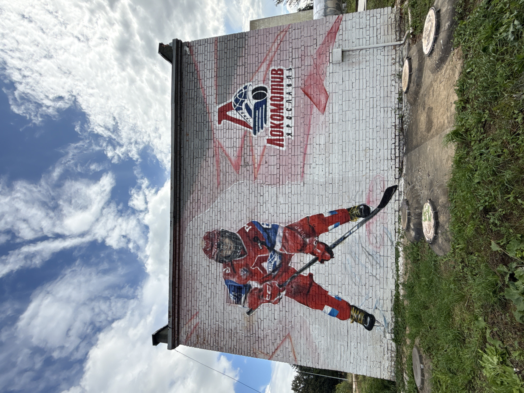 The image shows a mural on the side of a white brick building in Rusanovo, Russia. The mural depicts a hockey player in a red and blue uniform, holding a hockey stick. To the right of the player is the logo of the Lokomotiv Yaroslavl ice hockey team. The building is located in an outdoor environment with green grass and cloudy skies. There are several circular manhole covers visible in the foreground.