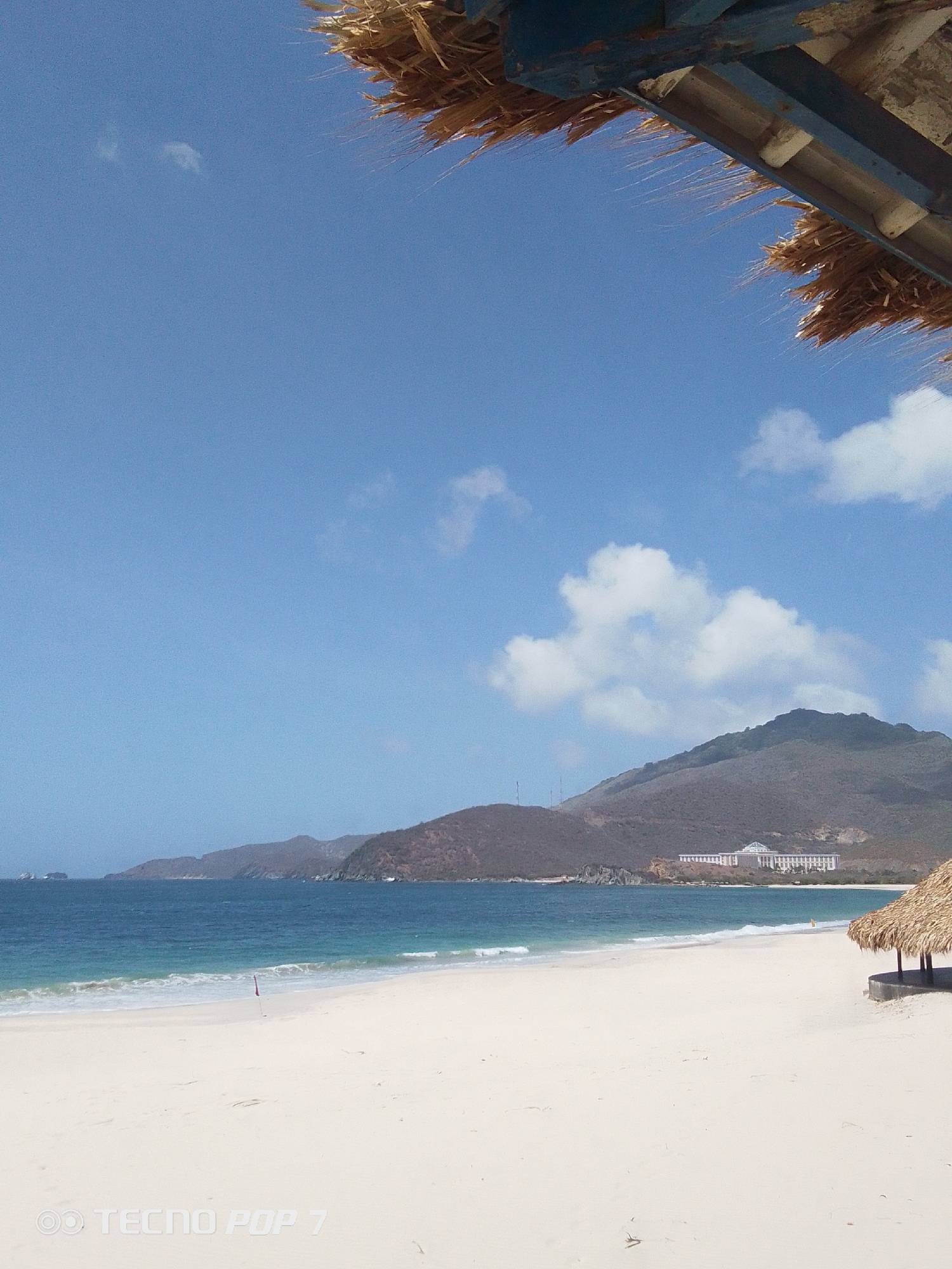 The image depicts a beach scene in Pedro González, Venezuela. The sky is a clear blue with scattered white clouds. The ocean is a turquoise color, and a white sandy beach stretches across the foreground. In the distance, mountains are visible. A building is situated on the coast. In the upper part of the image, a thatched roof structure is present. The beach appears deserted. The words "TECNO POP 7" are at the bottom of the image.