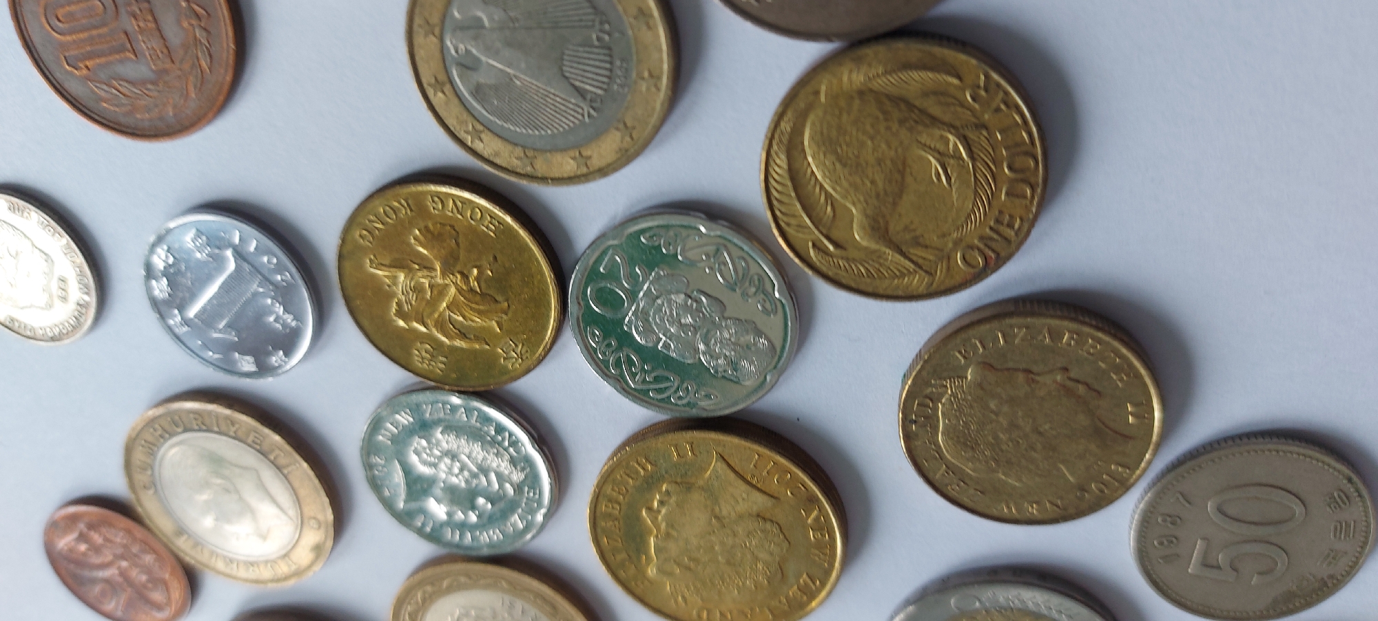 The image displays a collection of assorted coins arranged on a plain, light-colored surface. The coins vary in size, color, and design, suggesting they are from different countries and possibly different eras. Visible details include: a copper-colored coin with an embossed pattern on the left, a gold-colored coin with a profile of a person and text in a foreign language, a silver coin with a building-like design and "2011", a gold coin with a horse-like figure and "HONG KONG", a silver coin with a bird-like figure and "NEW ZEALAND", a gold coin with the profile of Queen Elizabeth II and "2013", a green and silver coin with intricate designs, a gold coin with a kiwi bird and "ONE DOLLAR", a gold coin with stars around the rim and an eagle design, and a silver coin with "1987" and "50" prominently displayed. There is no indication of the city of Chita, Russia, in the image. The scene is static, with no people or actions depicted.