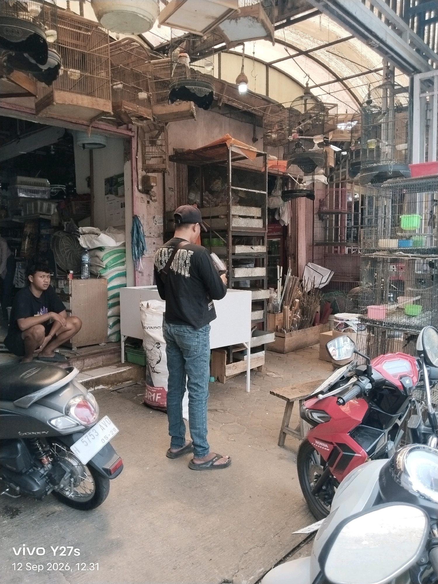 The image depicts an outdoor marketplace scene in Trimulyo, Indonesia. In the foreground, two motorcycles are parked. A silver scooter is on the left, and a red motorcycle is on the right. A young man with short, curly hair is crouched on the left side of the image, wearing a black t-shirt and shorts. He is looking towards the right. A man wearing a black t-shirt, blue jeans, and a black baseball cap is standing in the center of the image, facing away from the camera, and appears to be holding a small object, possibly a phone or a payment device. Behind the people and motorcycles, the marketplace is filled with various stalls. Numerous birdcages, some hanging and some on shelves, are visible, suggesting a bird market. Merchandise, including sacks of goods and what appear to be cleaning supplies or pet supplies, are stacked on shelves and tables. The environment appears to be a bustling marketplace with goods displayed openly.