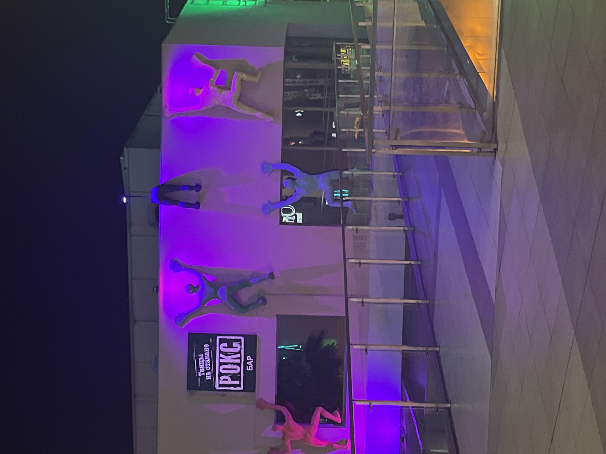 The image depicts a building with various neon-lit figures on its exterior, located in Sochi, Russia.
The building's facade is illuminated with purple and other colored lights, casting a glow on the surrounding area. The figures are stylized representations of human forms, seemingly engaged in various poses, possibly related to dancing. Below these figures is a sign that reads "РОКС БАР", with the phrase "танцы на стакане" written above it. This sign denotes the presence of a "Rocks Bar" with "dances on the glass" (likely pole dancing or similar) as its feature.
A staircase with glass panels and metal railings is visible near the building's facade. The scene appears to be taken at night, and the overall atmosphere is modern and somewhat stylized, indicative of an entertainment venue in a city like Sochi, which is known for its nightlife and tourist attractions. The background is dark, suggesting an urban setting at night.
