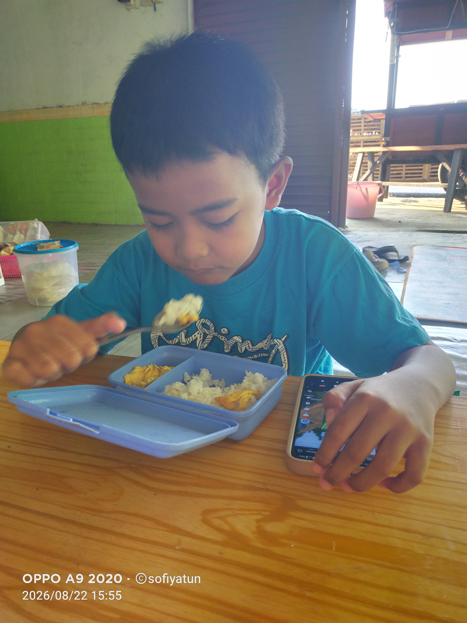 A young boy is eating lunch at a wooden table outdoors. He is wearing a blue t-shirt and holding a spoon with rice and scrambled eggs. A blue bento box with two compartments, one containing rice and the other scrambled eggs, sits in front of him. To his left is a clear plastic container with a blue lid, filled with white food. A mobile phone is on the table to his right. In the background, there is a green wall, a wooden structure, and bright sunlight. The lower portion of the image shows a watermark indicating the device used is an "OPPO A9 2020", the copyright holder is "@sofiyatun", and the date and time are "2026/08/22 15:55". The location is identified as Tayu, Indonesia.