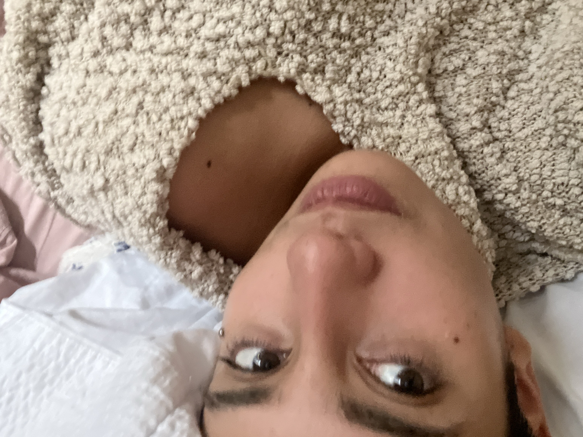 A close-up, rotated selfie shows a person with light skin wearing a textured, off-white sweater with a round neckline. The person has brown eyes, dark eyebrows, and pink lips. A small mole is visible on the left side of their chest, and another is on their left cheek. The background includes a white pillow, and a hint of a pink blanket or clothing item. No information regarding the location of the image, specifically Buenos Aires, Argentina, is present.