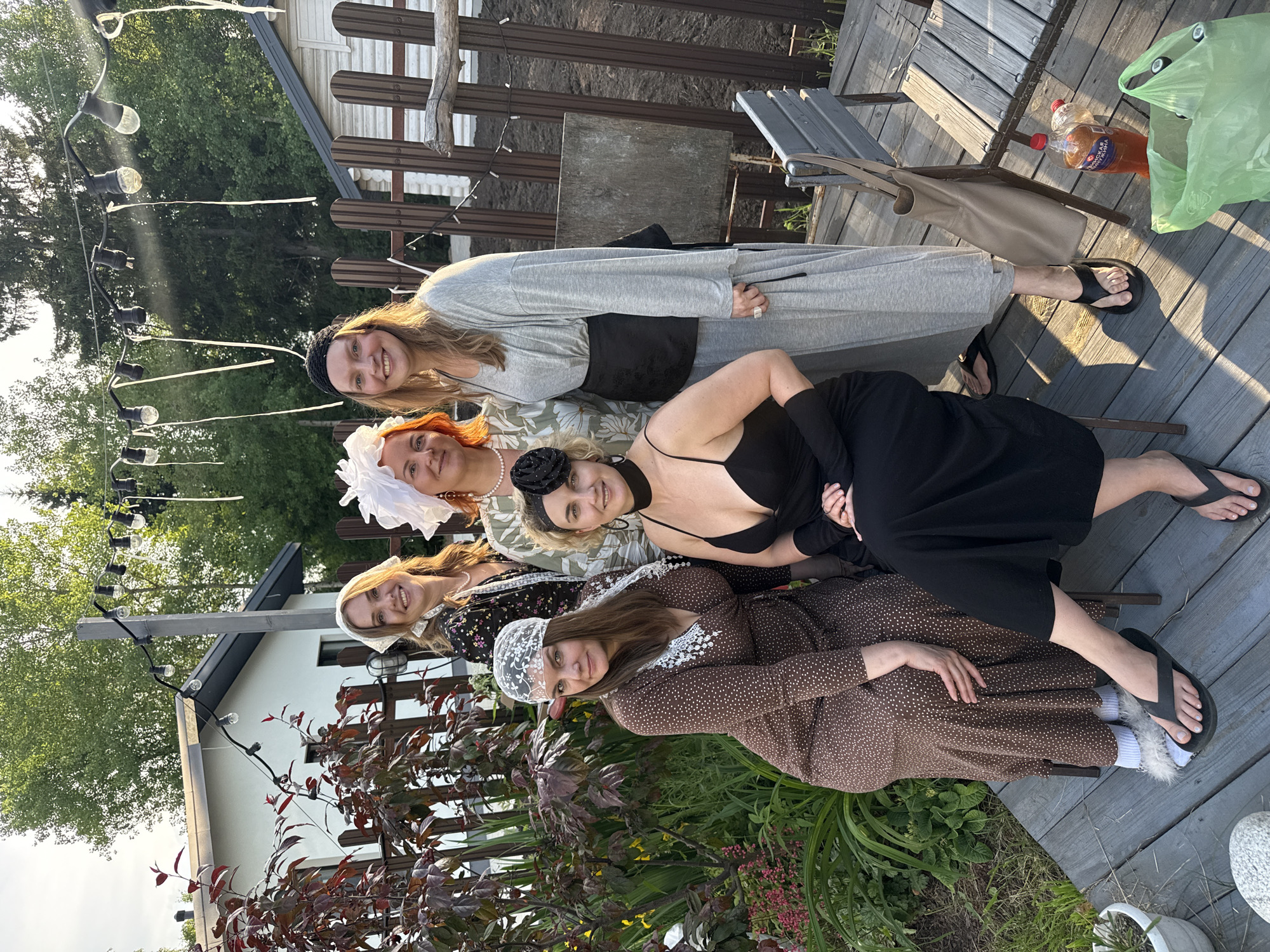 Six women are posing for a photo on a wooden deck in a backyard setting. The women are dressed in vintage-style clothing, including hats and dresses. They appear to be in their 30s and 40s. The background includes a white house with a dark roof, a wooden fence, and trees. String lights are hung across the yard. There are plants and flowers in the foreground. A table and chairs are also visible on the deck. The lighting suggests it is late afternoon or early evening. The presence of a white house with a dark roof and a wooden fence suggests a residential area, consistent with a backyard in Izhevsk, Russia.