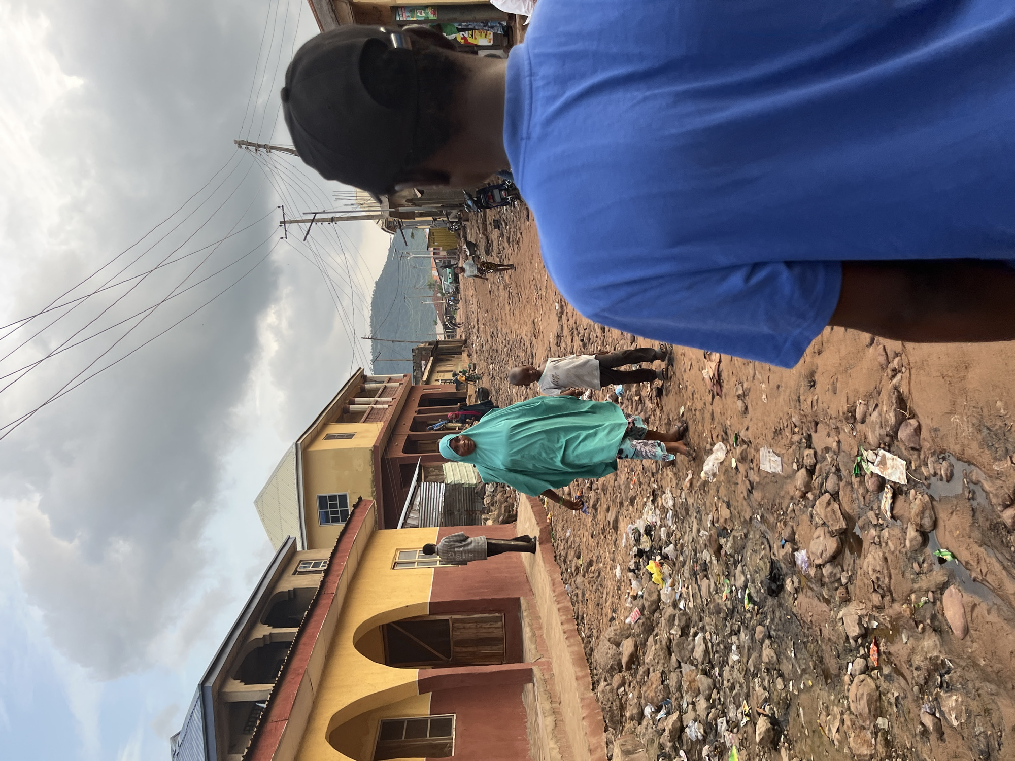 A medium shot captures a view from behind a person wearing a blue t-shirt and black baseball cap, looking down a dirt road in what appears to be a developing urban area. The road is littered with trash and debris, with puddles of murky water. On the left, a two-story building painted yellow and red with arched openings and balconies lines the street. Further down the road, other buildings with varying colors and architectural styles are visible, along with power lines and utility poles. Several people are walking on the road, including a woman in a teal hijab and a young child in a grey t-shirt and dark pants walking towards the camera. Another person in a plaid shirt is standing near the yellow building. The sky is overcast with heavy clouds. The overall scene depicts a busy, somewhat impoverished street in a town or city.