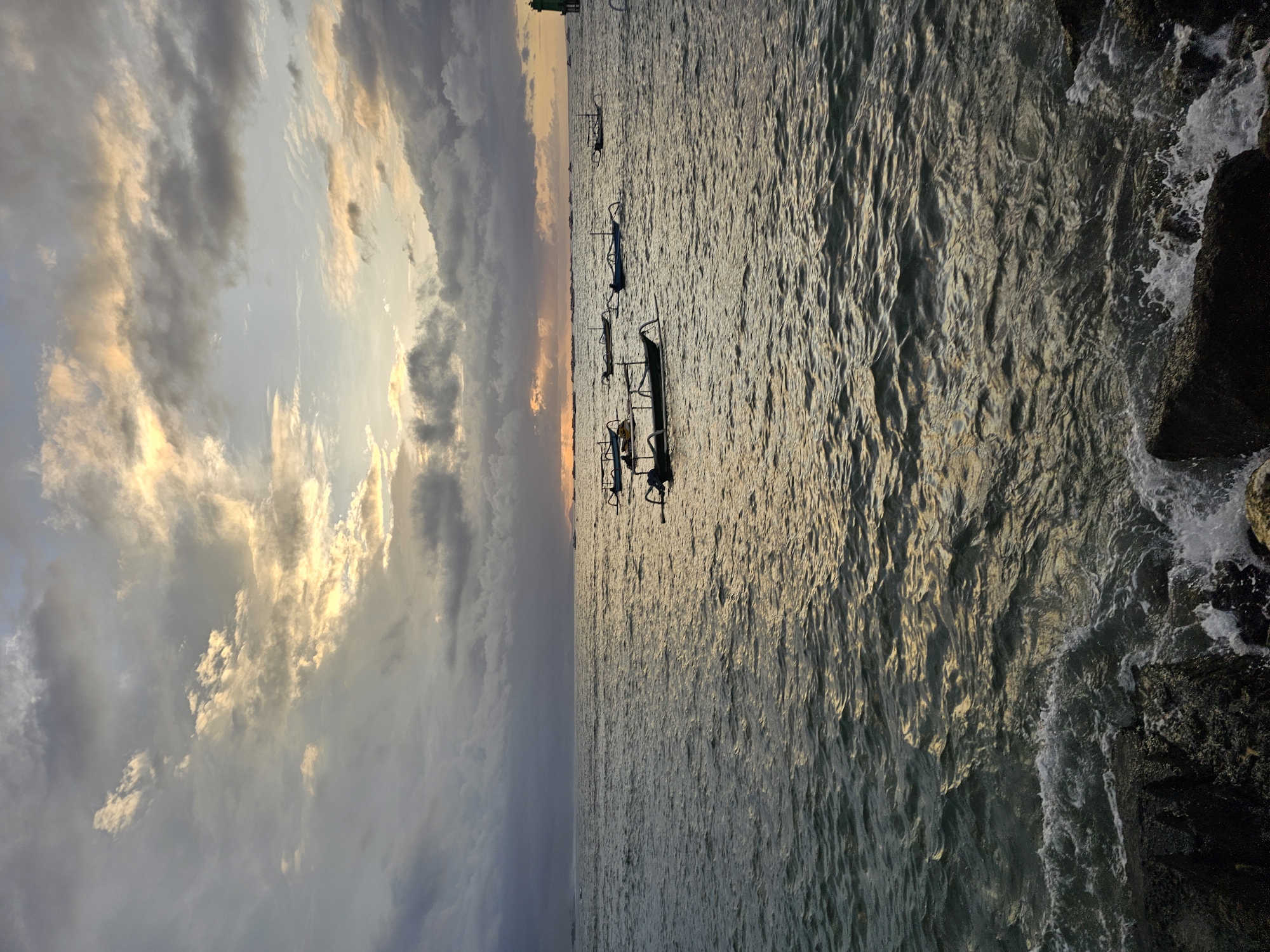 Captured in Denpasar, Indonesia, the image displays a serene seascape at dusk. Several traditional outrigger boats, possibly fishing vessels, are anchored in the gentle waves, silhouetted against the glowing sea. The waves create patterns of light on the water's surface. The photograph is vertically oriented, showing the clouds in the upper left and rocks on the right edge.