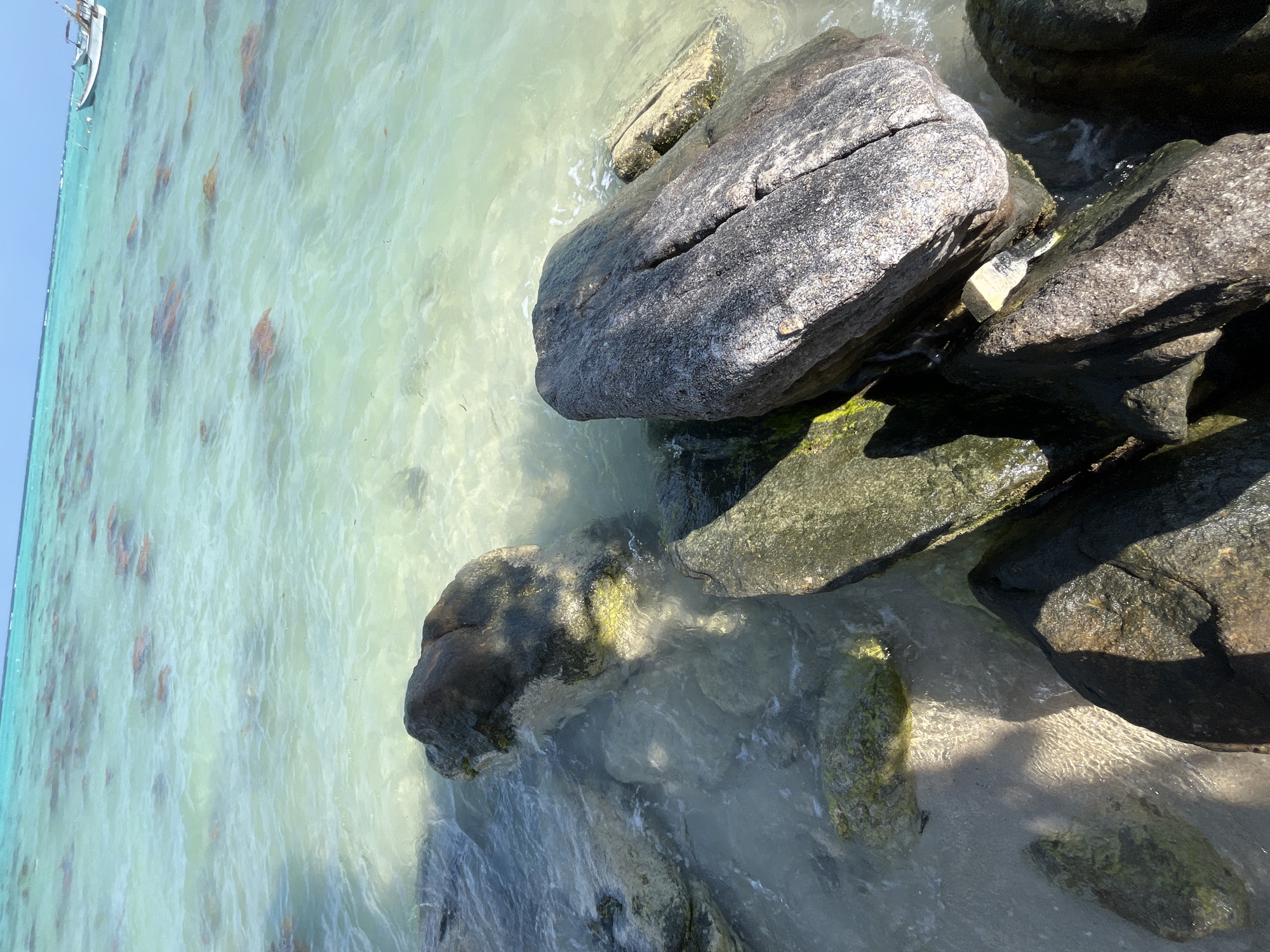 A high-angle, slightly tilted, medium shot captures a sunlit section of the ocean shore. Large, dark-colored rocks are scattered across the foreground, partially submerged in the shallow, crystal-clear turquoise water. The water's surface is disturbed by gentle ripples, revealing the sandy seabed beneath. In the distance, the water transitions to a deeper blue, and a small white boat with a brown canopy is visible on the horizon, as is a distant landmass. This scene is located at Legonipah, Indonesia.