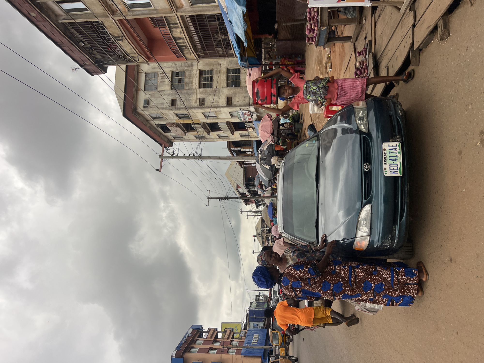In Nkpor, Nigeria, a street scene unfolds under a cloudy sky. A dark teal sedan, its license plate reading KED-417AE, is parked on a dirt and asphalt road, facing left. In front of the car, a young girl in a pink outfit balances a red gas cylinder on her head and a patterned bundle in her arms. To the left of the car, two women, one in a blue headscarf and a brown and blue patterned dress, and another in a patterned dress, stand near the vehicle. Further left, a person in an orange shirt and yellow shorts pushes a cart. The background features multi-story buildings with balconies and windows, some appearing weathered. A market stall with produce, possibly yams, is visible on the right. Umbrellas and other market structures dot the scene, indicating a bustling commercial area. Power lines crisscross the sky, and various vehicles, including a yellow tricycle, are present on the street.