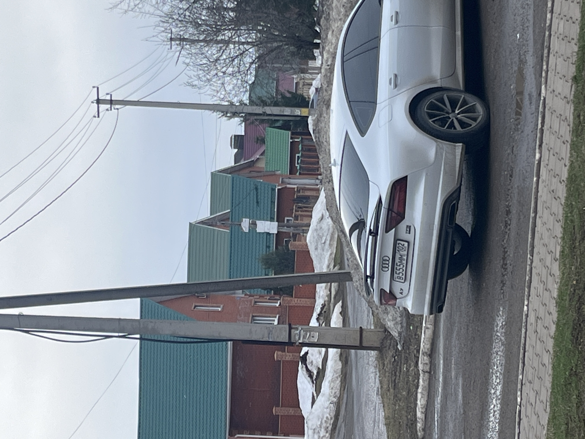 The image shows a white Audi car parked on a street in Neftekamsk, Russia. The car's license plate is B555MM 102. Houses with green and red roofs are visible in the background. A utility pole with power lines stands nearby. Patches of snow are present on the ground. The street appears wet, likely due to melted snow. The image is taken from a slightly elevated angle.