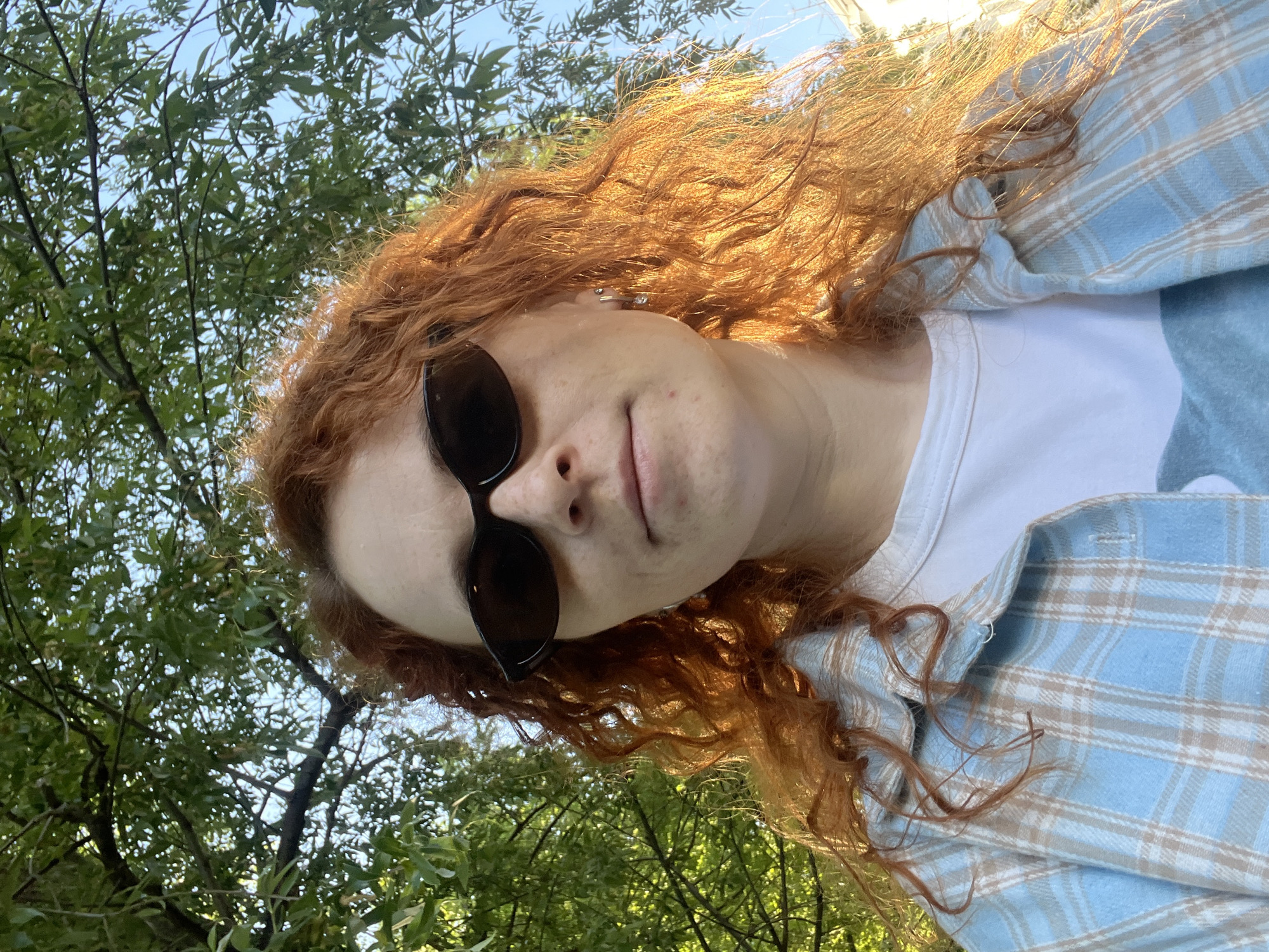 A woman with auburn hair and sunglasses poses for a selfie in an outdoor setting with trees visible in the background. She wears a white t-shirt and a light blue plaid flannel shirt. The image is taken from a low angle. The exact location in Chaikovsky, Russia cannot be determined from the image alone.