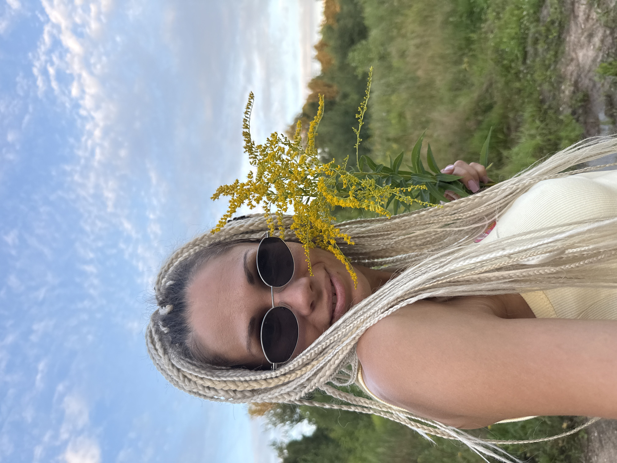 A young woman with blonde braided hair and sunglasses holds a sprig of yellow flowers. She is wearing a yellow tank top and is in a grassy outdoor setting. The background consists of green trees and a cloudy blue sky. There is no information in the image to indicate the location is Yaroslavl, Russia.