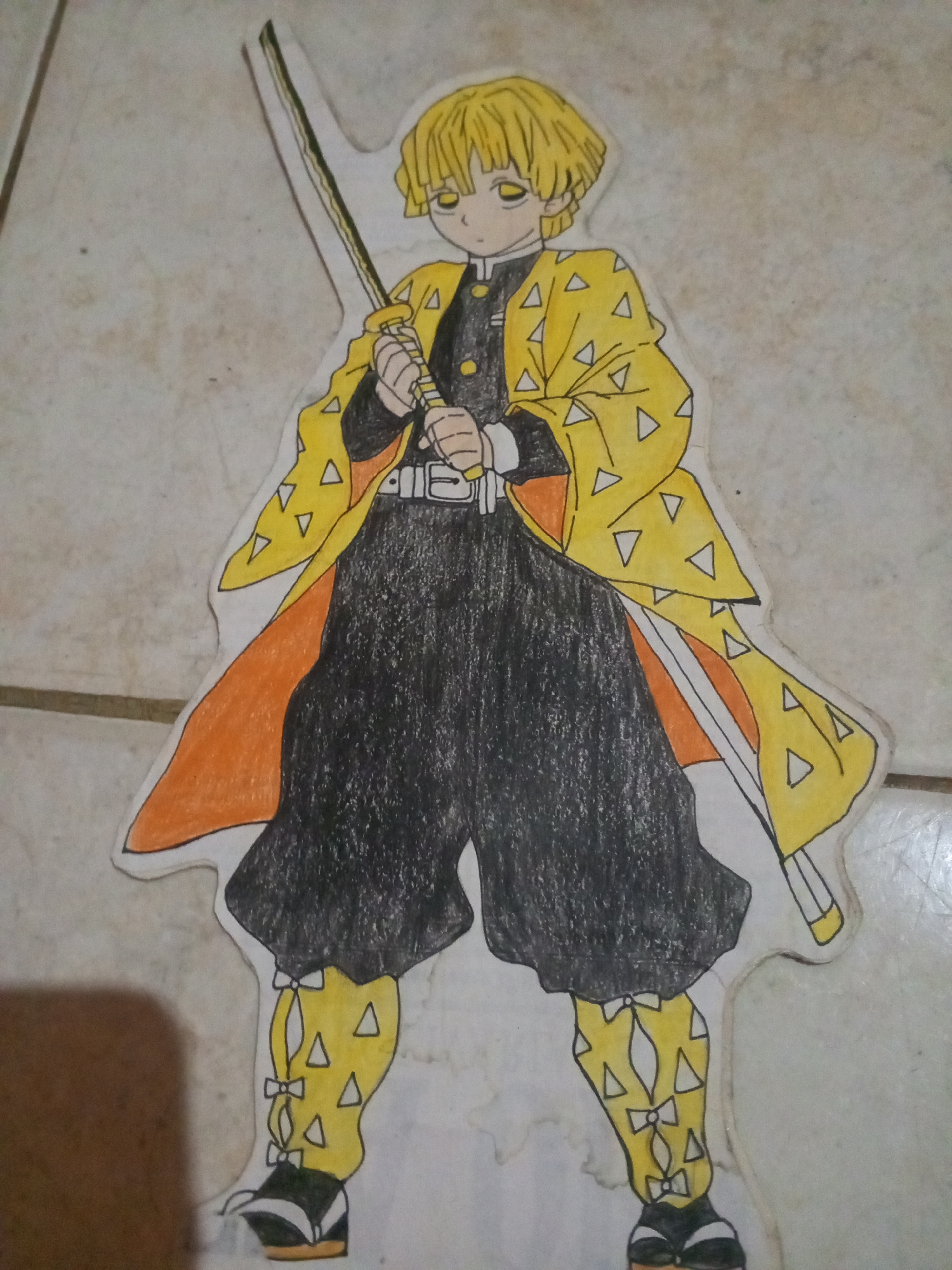 A drawing of an anime character, Zenitsu Agatsuma from Demon Slayer, is placed on a tiled floor. The drawing depicts Zenitsu standing with a sword in his hand, wearing his signature yellow haori with triangular patterns, black uniform, and striped socks. The background is a light-colored, speckled tile floor, typical of household environments. There is no indication that this scene is located in Altagracia, Venezuela. The image focuses solely on the drawing and its immediate surroundings on the floor.