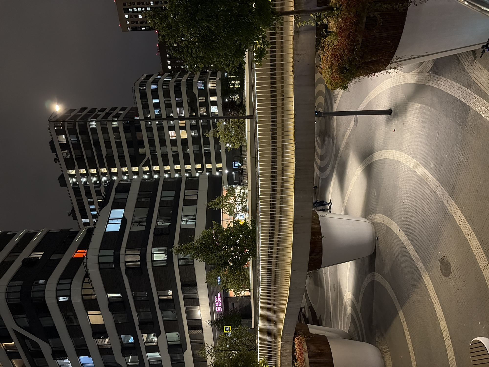 This is a nighttime exterior shot of the Krylatsky Hills residential complex in Moscow, Russia. The image captures the modern architecture of the buildings, characterized by their dark facades and numerous illuminated windows. A pedestrian bridge with a lit railing spans across the foreground, casting light onto the patterned pavement below. Large white planters, some containing autumn foliage, are strategically placed. The scene is illuminated by artificial lights from the buildings and street lamps, creating a somewhat moody atmosphere under a dark sky. Two individuals are visible walking on the pavement beneath the bridge. The overall impression is of a contemporary urban environment at night.