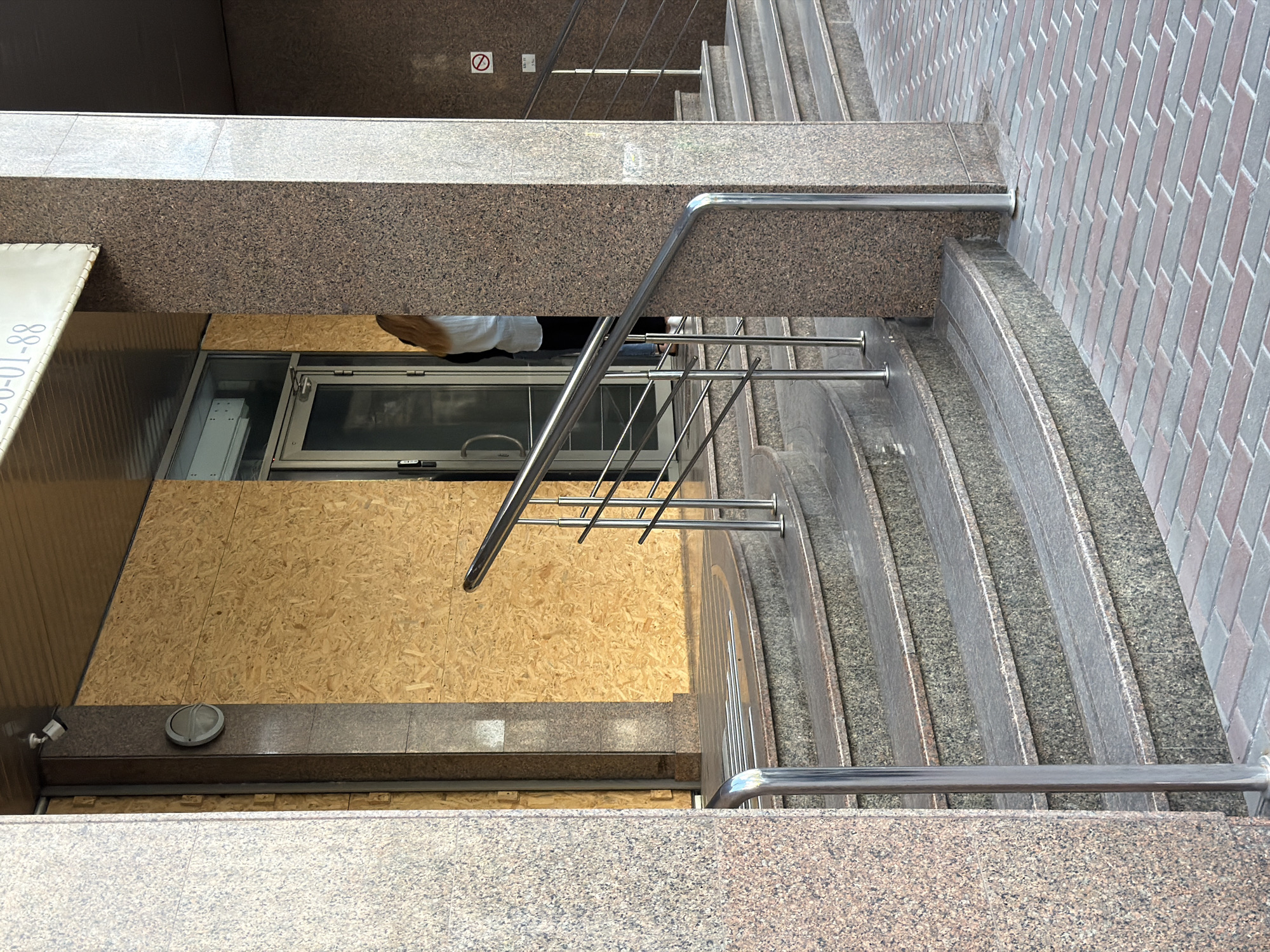 The image depicts the entrance to a building in Kyiv, Ukraine, which has been secured with plywood. The plywood is covering the entire storefront, suggesting damage or an attempt to protect the interior. A glass door is visible behind the plywood, and a person, appearing to be a woman with blonde hair, is partially visible standing just inside the doorway. There are also a set of granite stairs leading up to the entrance, with a metal handrail. The ground in front of the stairs is paved with brick. The overall scene suggests a situation of disruption or preparation for potential harm.