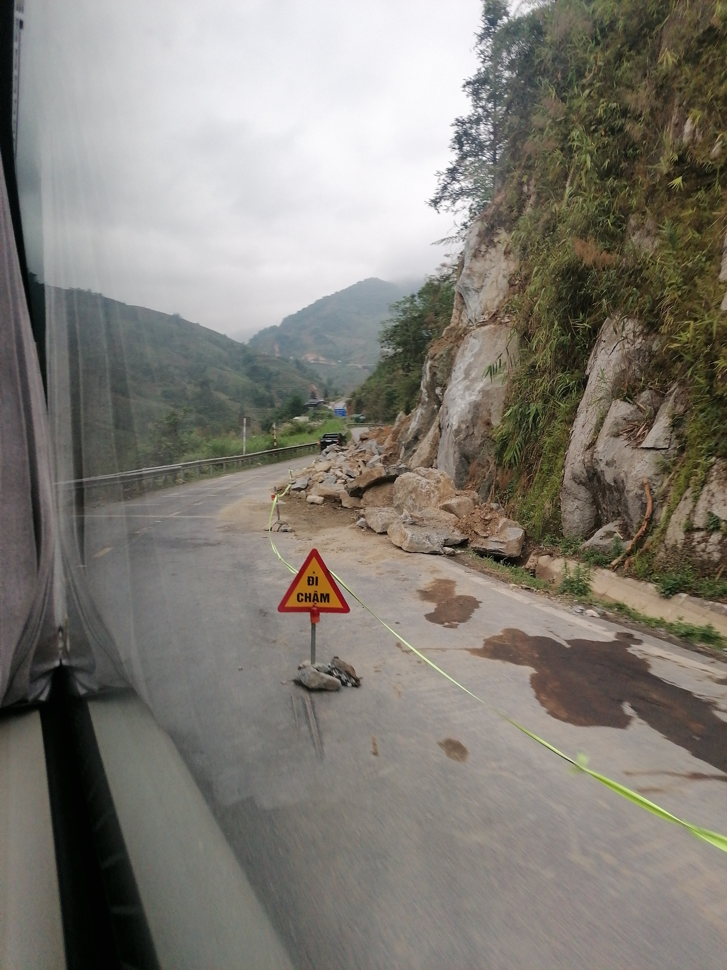 The image shows a road in an area of Unknown, Unknown that has been partially blocked by a rockslide. A red and yellow triangular sign with the text "DI CHAM" is present, likely serving as a warning to drivers. A green ribbon is strung across the road as a barrier. The landslide is positioned next to a rocky, vegetated cliff. The road appears wet and there are mountains visible in the background under an overcast sky. The image seems to have been taken from inside a vehicle, as part of the window is visible.