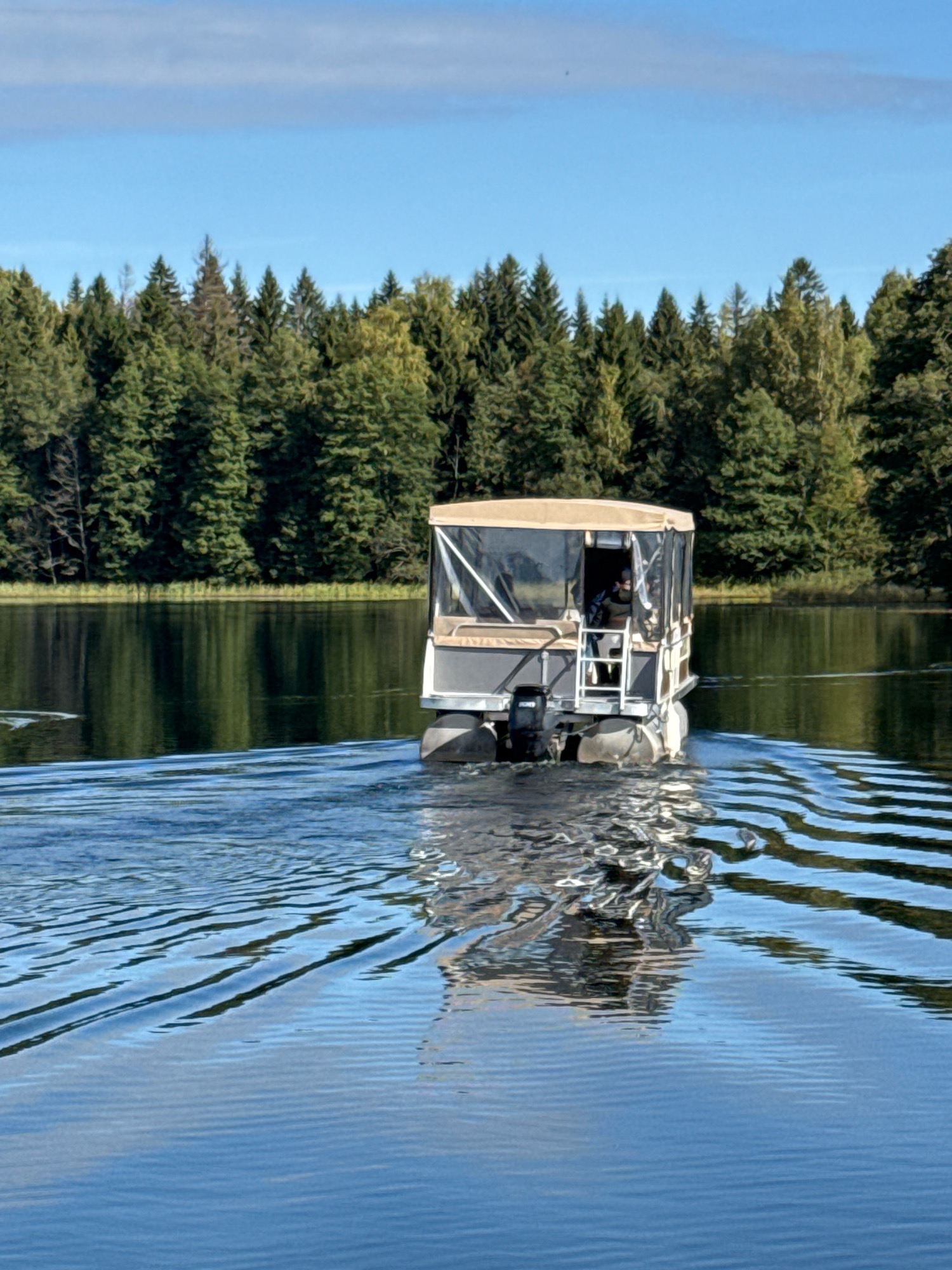 An aerial rear view shows a pontoon boat moving across a calm lake with a dense forest lining the shore. The boat has a tan canopy and a silver cabin with a person visible inside, operating the vessel. The water is blue and slightly rippled, with reflections of the sky and trees visible. The sky is clear blue with a few wispy clouds. The overall scene depicts a serene outdoor environment, likely in a rural or natural setting outside of a city.