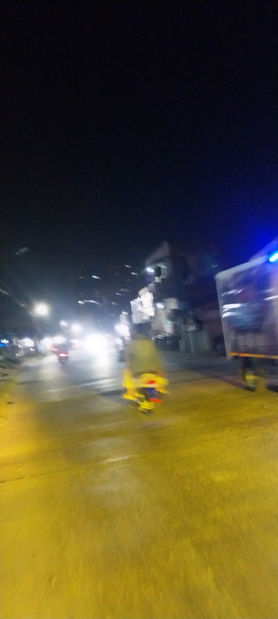 The image depicts a blurred nighttime scene on a street in Kelapa Dua, Indonesia. A yellow scooter or motorcycle with a rider is prominently featured in the center, moving away from the viewer. To the right, a portion of a truck, possibly a delivery vehicle, is visible with blue lights illuminated on its roof. The street is wet and reflects the bright headlights of other vehicles and streetlights, creating streaks of light across the yellow-tinted road surface. In the background, the indistinct shapes of buildings and other vehicles can be observed, suggesting an urban environment. The overall impression is one of motion and a busy, illuminated streetscape at night.