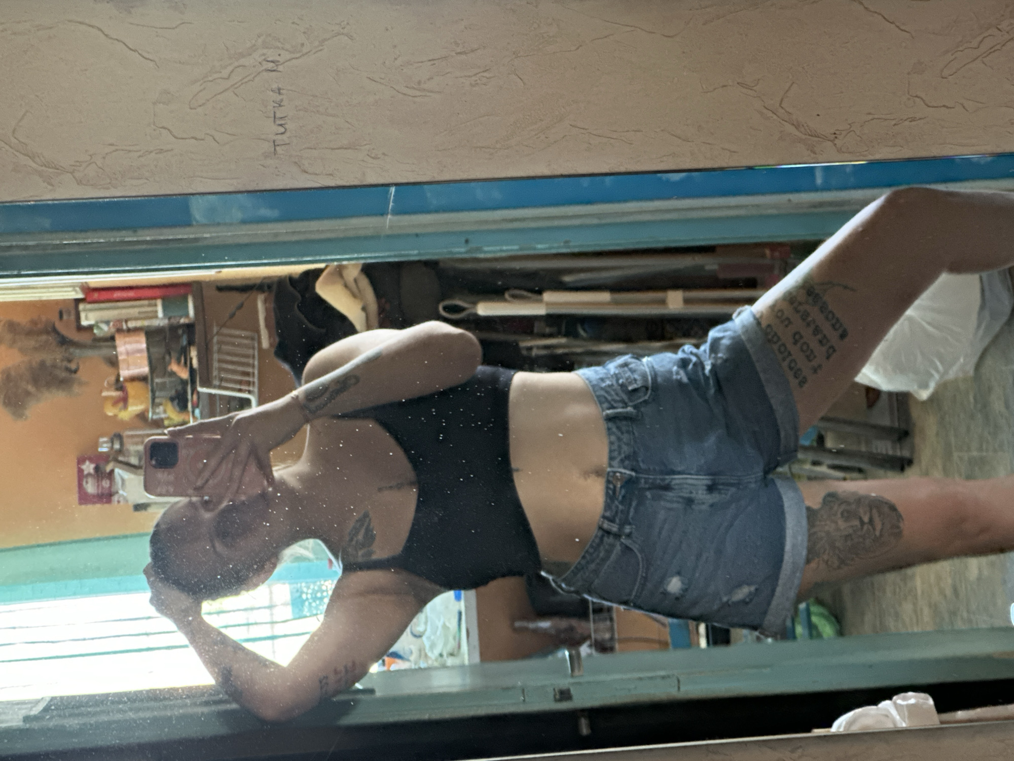 A woman is taking a mirror selfie in what appears to be a bedroom or living space. She is wearing a black tank top and denim shorts, with tattoos visible on her arms and legs. Her hair is pulled back, and she is holding a pink smartphone in her right hand, capturing the reflection. The background is cluttered with various objects, including shelves, clothing, and a window with vertical blinds. The wall behind her has a textured finish and some writing. The overall impression is of a casual, everyday scene. It is not possible to determine the specific location in Plovdiv, Bulgaria, based on the visual information provided in the image.