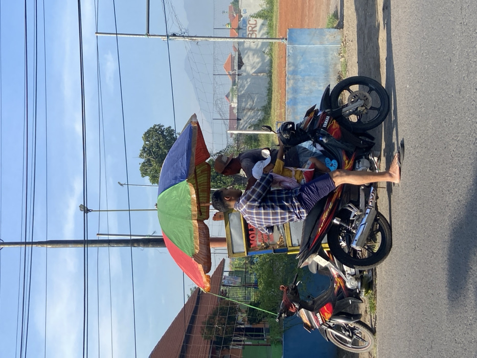 The image displays an outdoor street scene in Balongdowo, Indonesia, under a clear blue sky during daylight. In the foreground, a male adult customer, wearing a blue and white plaid shirt and dark shorts, is seated barefoot on a black Yamaha motorcycle with red and orange flame decals. A young child, dressed in a white hat, yellow shirt, and pink backpack, sits directly in front of the adult on the same motorcycle. Both face a male vendor, who wears a hat and dark shirt, standing behind a mobile food cart positioned under a large, multi-colored umbrella featuring red, green, and blue segments. The cart is light-colored and exhibits partial text, including "MOLE" and "GORENG." Another red and black Yamaha motorcycle is parked on the left side. The background features a dark utility pole with multiple overhead wires, a brown-roofed building behind a chain-link fence, and a blue-painted concrete barrier with "BRC" inscribed on it. Beyond the barrier lies an open reddish-brown dirt field, with trees and distant mountains visible against the horizon.