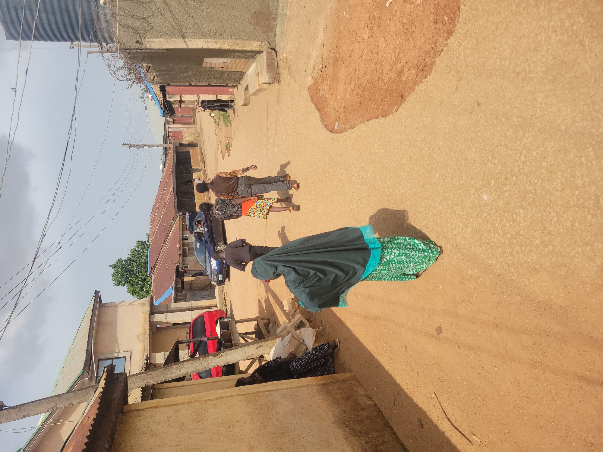 The image depicts a street scene in Jos, Nigeria. The road is unpaved and a reddish-brown color. Several people are present. Two men are walking in the same direction, while a woman in a green and dark-colored outfit is walking in a different direction. A person appears to be lying on the steps of a building. A blue car is parked on the side of the road. The buildings appear to be constructed from concrete. Above them, several electrical wires are present.