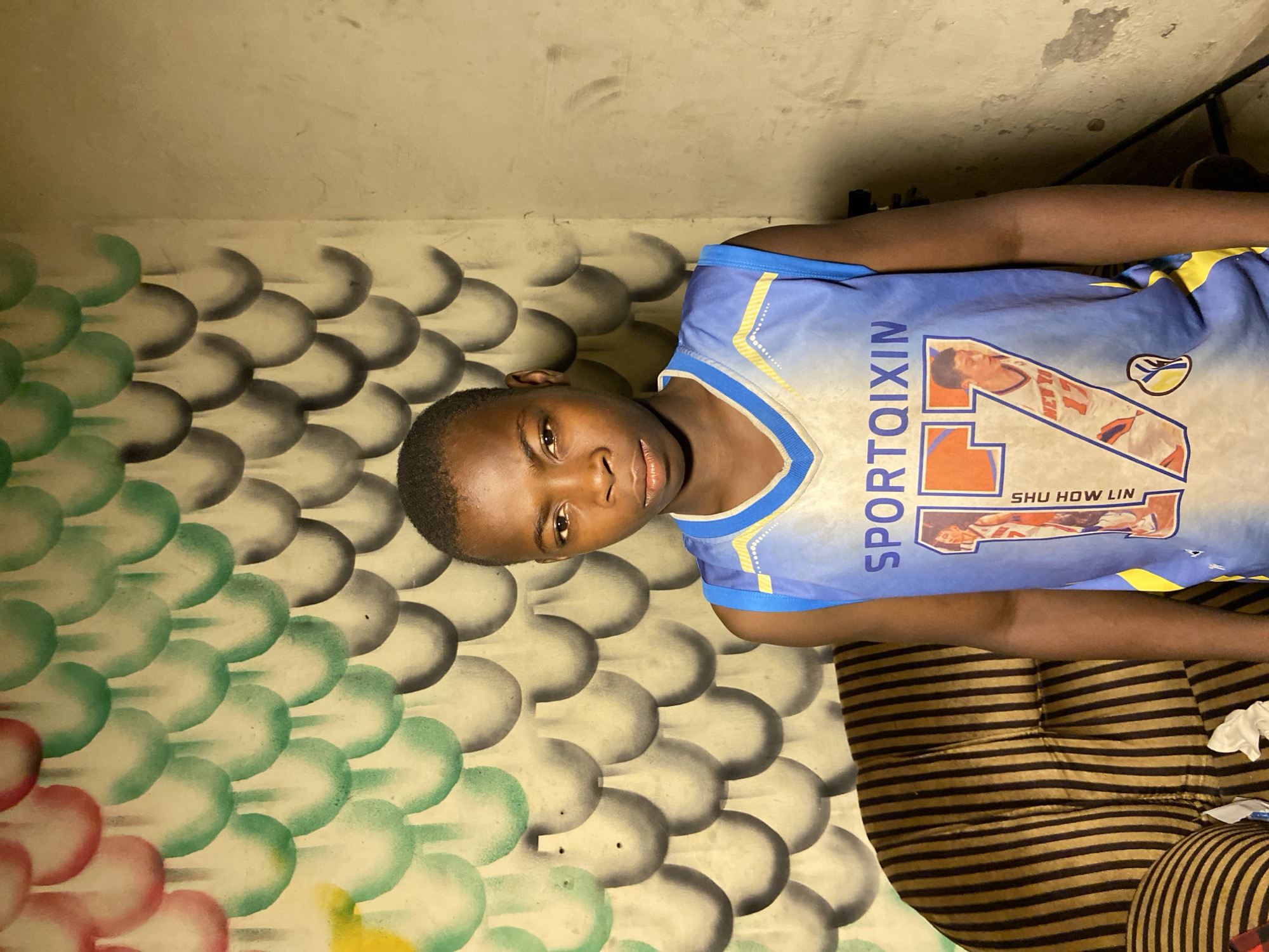 A young, dark-skinned boy, likely between 10 and 14 years of age, is positioned indoors. He is wearing a blue and yellow basketball jersey with the number 17, the name "SPORTQIXIN" vertically printed on the left, and the name "SHU HOW LIN" written under a player graphic on the front. The boy is looking at the camera with a direct gaze.
The backdrop consists of a wall with a painted bubble or scale pattern in varying shades of green, grey, and red near the bottom. Above the pattern, the wall is a dull, light beige color with a visible patch. To the right, near the top of the frame, there is a glimpse of what appears to be a dark metal or wooden structure.
The boy is sitting on or near a striped surface with dark brown and tan stripes. Some crumpled white paper or fabric is also present. The overall lighting is subdued and appears to be artificial. The location is Mararaba, Nigeria.