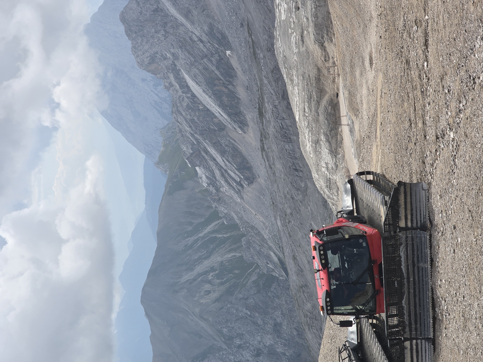 A red snow groomer is parked on a rocky, barren mountain slope. The vehicle has a large metal blade in the front and tracks on the bottom. In the background, there are multiple layers of mountains with steep, rocky faces, receding into a hazy, blue distance. The sky is partly cloudy with large, white clouds. The scene appears to be a high-altitude, mountainous environment, likely in the Bavarian Alps near Garmisch-Partenkirchen, Germany, given the type of vehicle and landscape.