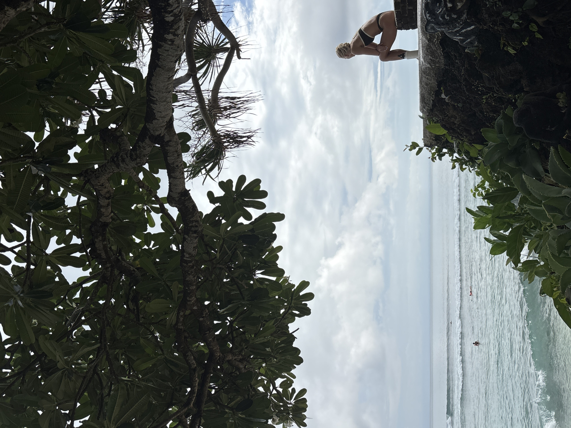 A horizontal view of a coastal scene in Kuta Selatan, Indonesia. A person in a black swimsuit crouches on the edge of a low, dark-colored wall. Below, the ocean stretches to the horizon, with gentle waves and several people swimming. The view is partially obscured by trees with dense green foliage on the left and right. The sky is overcast with light gray clouds.