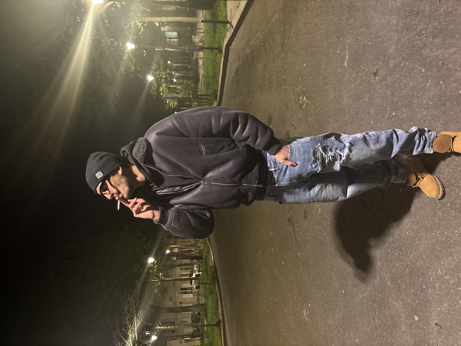 A male individual stands centrally in a paved street at night in Paris, France. He wears a black beanie with a visible emblem, dark-rimmed glasses, a dark grey or black bomber jacket with a fleece-lined collar over a dark hoodie, light blue distressed jeans with tears on the left thigh, and tan work boots. His right hand holds a lit cigarette near his mouth, while his left hand rests in his left jeans pocket. He looks upwards and to his right, away from the camera. The street is illuminated by multiple traditional Parisian streetlights with distinctive glowing lamps, casting light rays and shadows, including the subject's shadow to his left. The background features tree-lined sidewalks or verges with grass, and beyond them, multi-story residential buildings are visible on the left. A white car is parked in the distance on the left, and a red circular traffic sign is also present on the left side of the street. The overall architecture and street furniture are consistent with a Parisian urban environment.